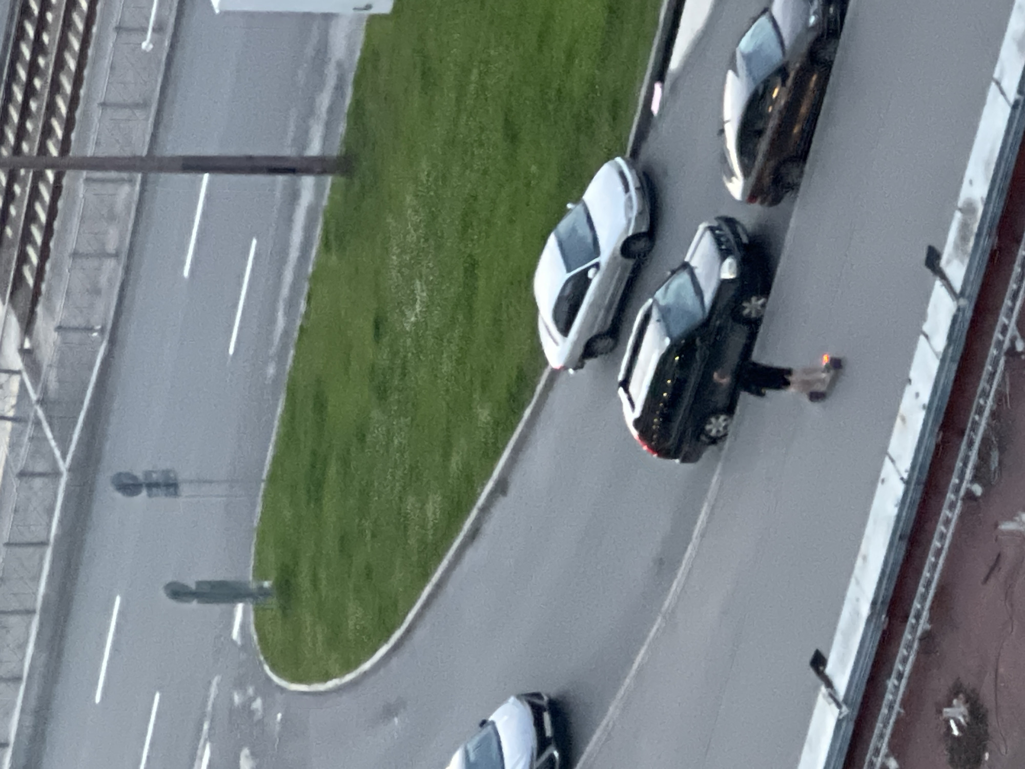 A low-angle, slightly blurry, outdoor shot shows a man on a scooter on a wet asphalt road, in the city of an unknown location in Russia. The man is wearing dark shorts and a black shirt, and is moving from left to right. Behind him, a black SUV is visible, followed by a brown car. To the left of the SUV, a silver sedan is turning a corner. In the background, a green grassy area is visible, with two traffic signs. Further in the background, train tracks are visible.