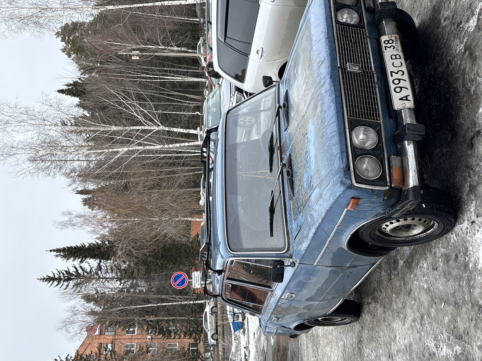 The image depicts a blue Lada car parked on an icy street in Ust-Ilimsk, Russia. The license plate reads "A993CB 38 RUS." A "No Stopping" sign is visible nearby. The background shows a brick building and bare trees under a gray sky. Other cars are parked nearby. The scene suggests a cold, possibly wintery urban environment.