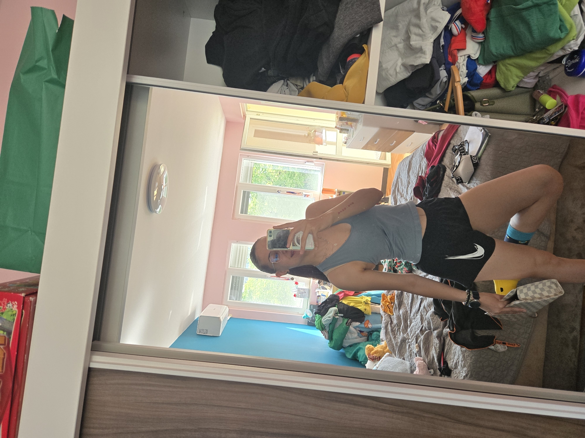 The image depicts a person reflected in a mirror. The person is holding a phone and appears to be taking a selfie. The reflection reveals a cluttered room with a bed, clothes, and other items scattered around. The room has pink walls and a blue floor. A window is visible, suggesting it is daytime. Based on the provided information, the location is in the city of Pleven, Bulgaria.