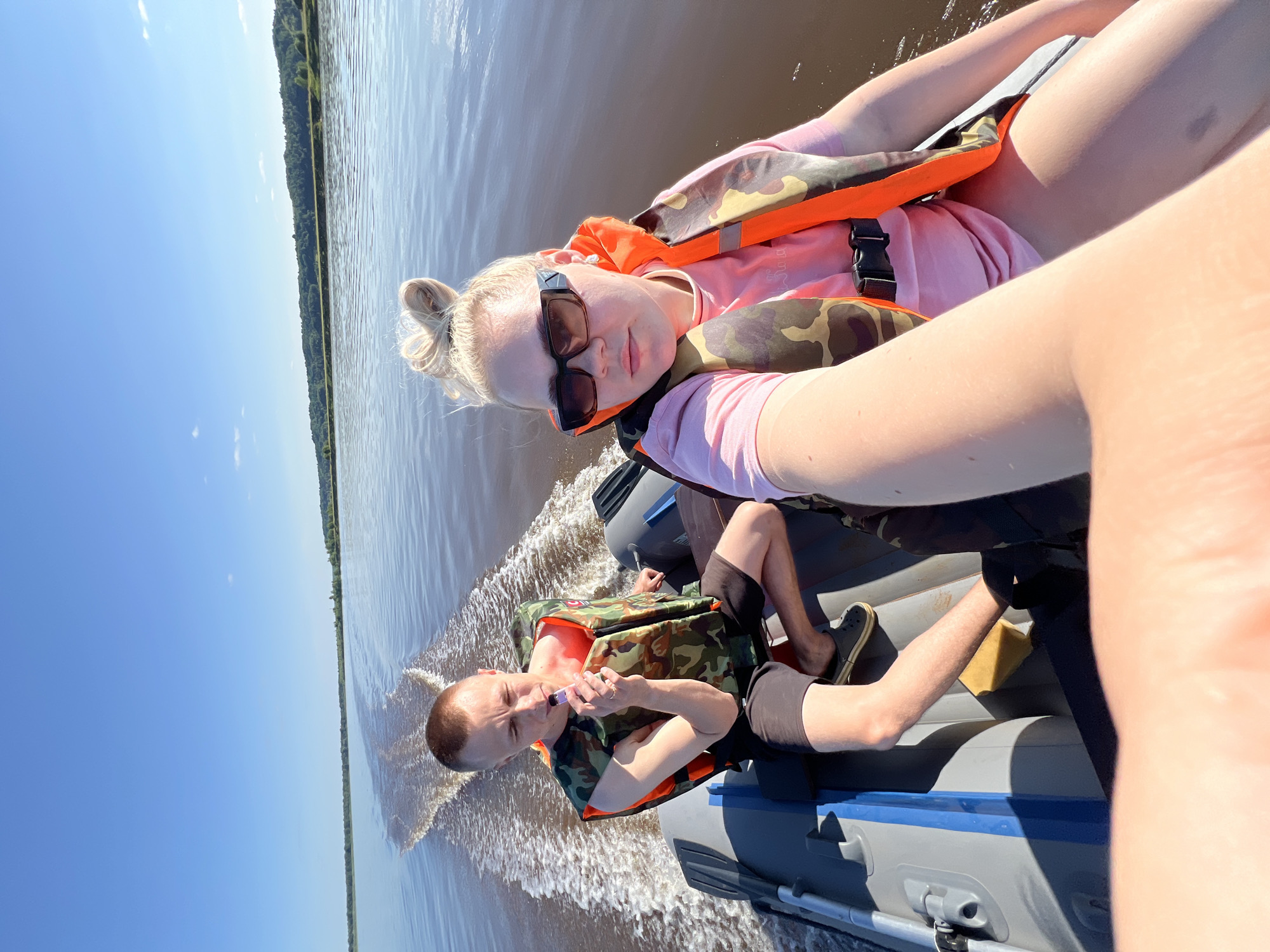 Two individuals, a man and a woman, are seated in an inflatable boat on a body of water, likely a river. The man, on the left, is wearing a camouflage life vest and dark shorts, and is holding a vaping device to his mouth. The woman, on the right, is wearing a pink t-shirt, sunglasses, and a similar life vest. She appears to be taking a selfie. The boat is moving, leaving a wake behind it. The environment is natural, with a clear blue sky and a treeline visible in the distance. The image was taken on a sunny day. There is no indication of the city of Mari-Malmyzh, Russia in the image.