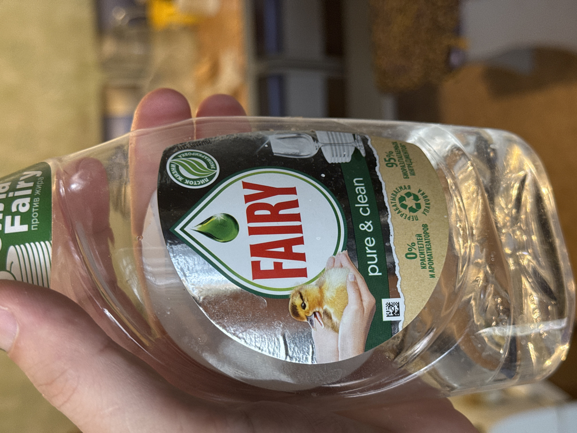 A hand holds a transparent plastic bottle of Fairy brand dish soap. The label reads "FAIRY" in large red letters with "pure & clean" underneath. A graphic shows a hand holding a small duckling. The label also includes green text and the words "95% биоразлагаемых ингредиентов" in Russian, indicating 95% biodegradable ingredients. The bottle is partially filled with clear liquid. The background is blurred and indistinct. There is no information in the image about the location of Chuprino, Russia.