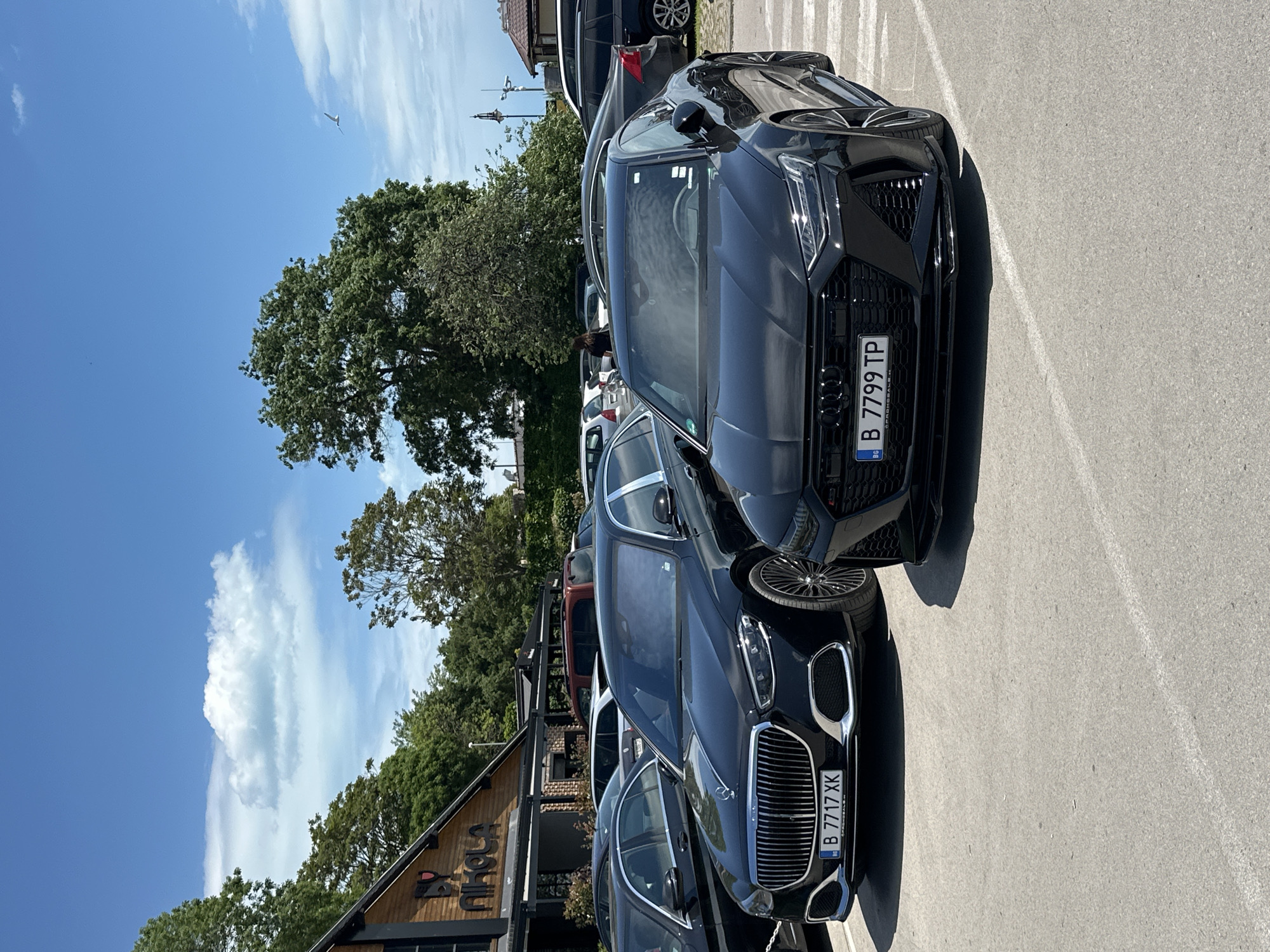 The image shows a parking area in Varna, Bulgaria, featuring two black luxury cars: a BMW and an Audi. The BMW's license plate reads "B 7717 XK," while the Audi's license plate is "B 7799 TP." Both vehicles are parked closely together. Behind the BMW, there is a wooden building with the word "Nikola" visible. In the background, there is also a view of trees and a blue sky with clouds. People are visible further in the background, near the trees, but they are not a focal point of the image. The ground is paved with light-colored concrete.