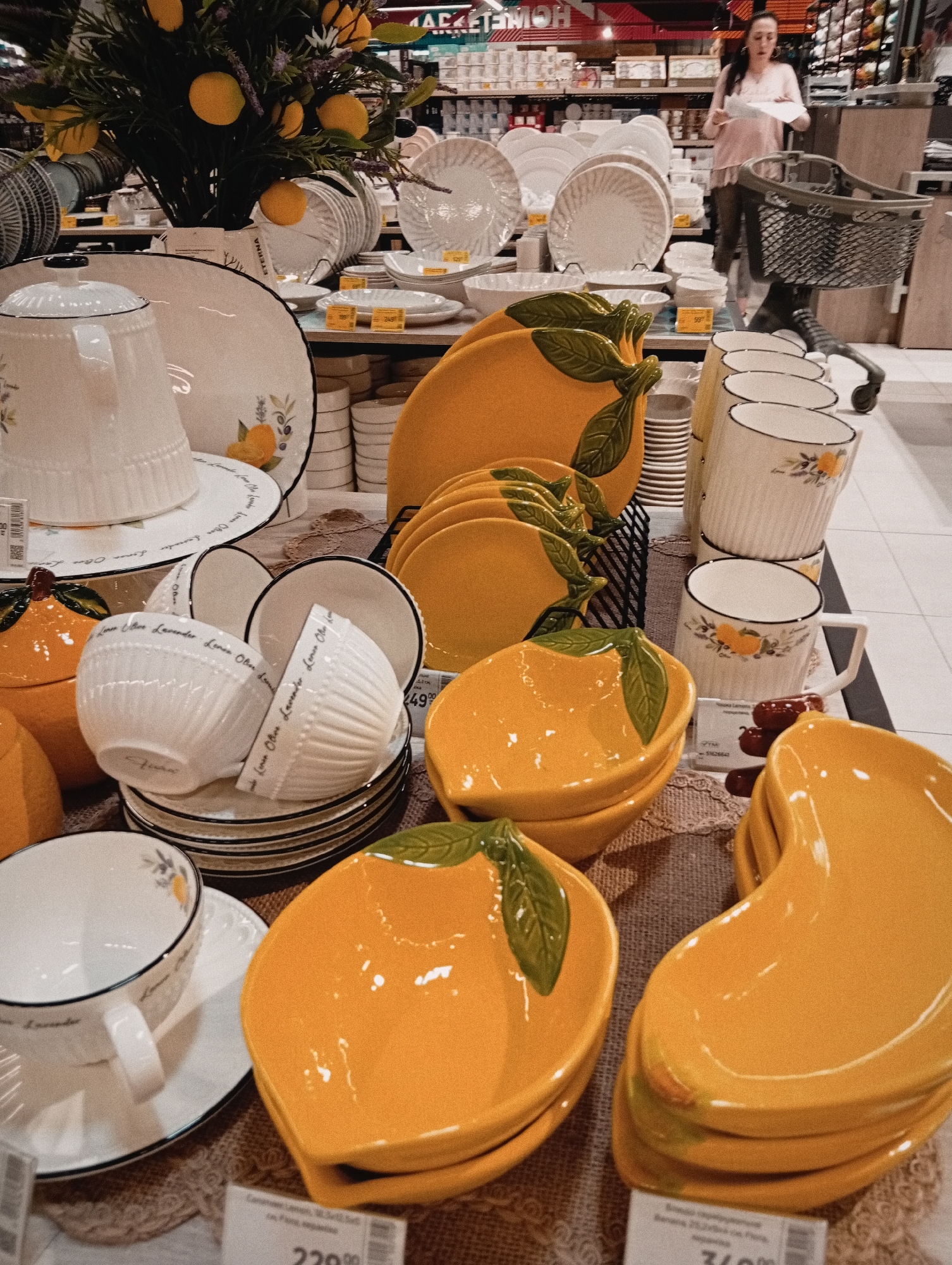 Here's a detailed description of the image:
**General Overview:**
The image appears to be taken inside a large retail store, possibly a home goods or department store. There are displays of dishes and tableware. The setting suggests a commercial environment.
**Objects:**
- Stacks of plates, bowls, cups, and saucers in various designs.
- Prominent are items with a lemon or citrus theme, including yellow lemon-shaped serving dishes and tableware decorated with lemons.
- Some of the tableware features a lavender motif as well.
- Prices tags are visible, indicating the items are for sale.
- There's a decorative arrangement with faux lemons and flowers.
- A shopping cart is partially visible in the background.
**People:**
- A woman with her hair tied back is visible pushing a shopping cart near the back of the frame. She is looking at something in her hands.
**Environment:**
- The background is somewhat blurry, suggesting depth of field. It shows shelves stocked with various items, characteristic of a retail store.
- The lighting appears to be artificial, typical of a store setting.
- The image suggests a clean, well-organized shopping environment.
**Actions/Interactions:**
- It is reasonable to assume that the woman in the background is shopping.
- The presence of price tags indicates that these items are for sale.
**Location:**
- Based on the visible OCR text and information embedded in the image, the location is Poltava, Ukraine.