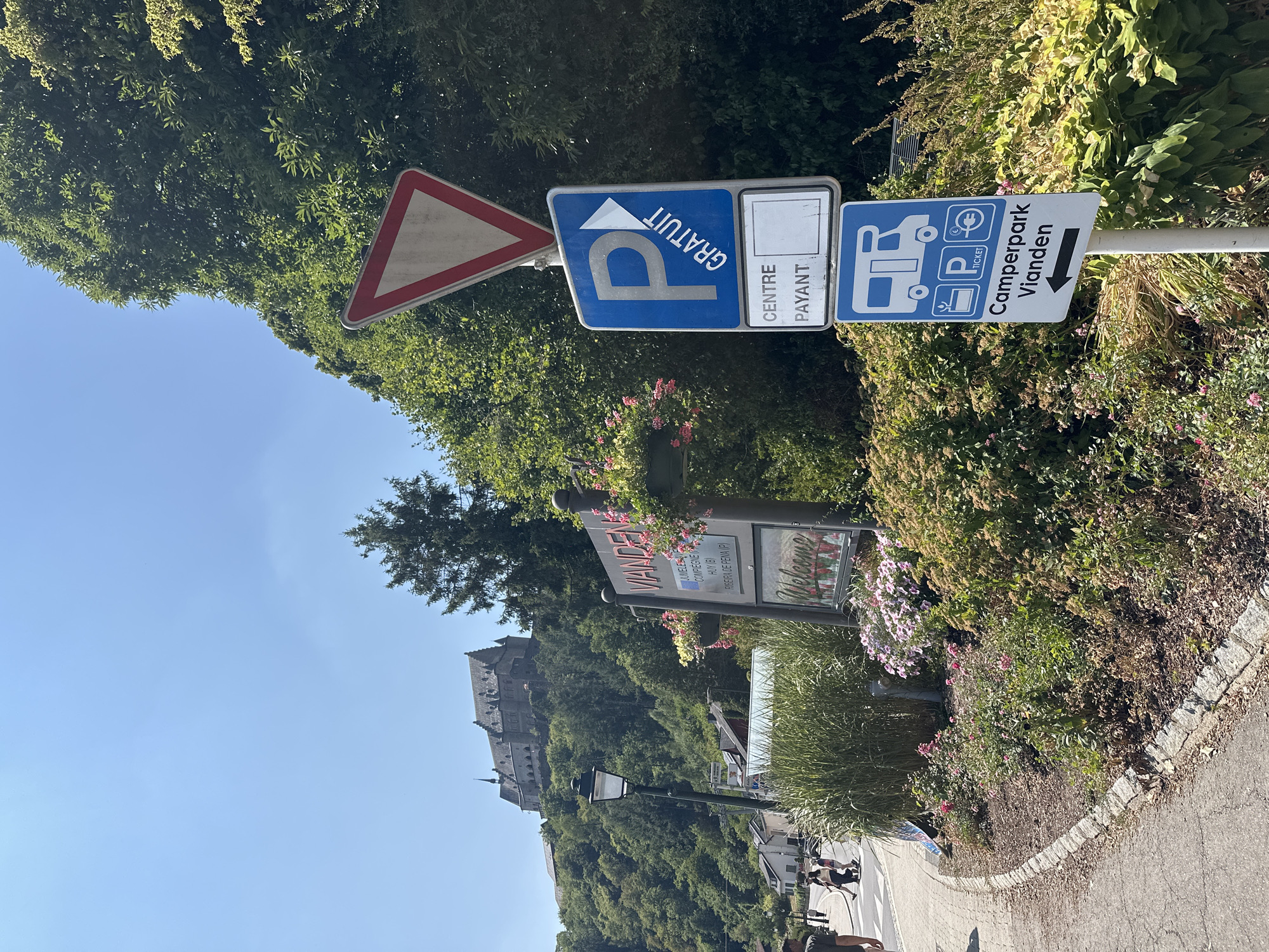 The image captures a sunny daytime scene in Vianden, Luxembourg. In the background, the prominent Vianden Castle sits atop a lush green hill. In the foreground, a street sign indicates "VIANDEN" with a list of cities including Jumel(B), Compeigne (F), Huy (B), and Ribeira de Pena (P). A "Welcome" sign with floral accents is also visible.
Several road signs are positioned on the right side of the image. The topmost sign is a red and white yield sign. Below it, a blue sign with a white "P" and an arrow points to the right, indicating "GRATUIT" (free parking). Underneath this, a sign reads "CENTRE PAYANT," suggesting paid parking in the town center, with a blank space below it. The lowest sign displays an icon of a camper van, a plant, a parking ticket symbol, and a euro symbol, with text reading "Camperpark Vianden" and an arrow pointing left.
A paved sidewalk lines the left side of the image, with a few individuals walking. Lush greenery, including tall grasses, bushes, and flowers, surrounds the signs and street area, with a lamp post also present. The environment appears to be a scenic tourist area.