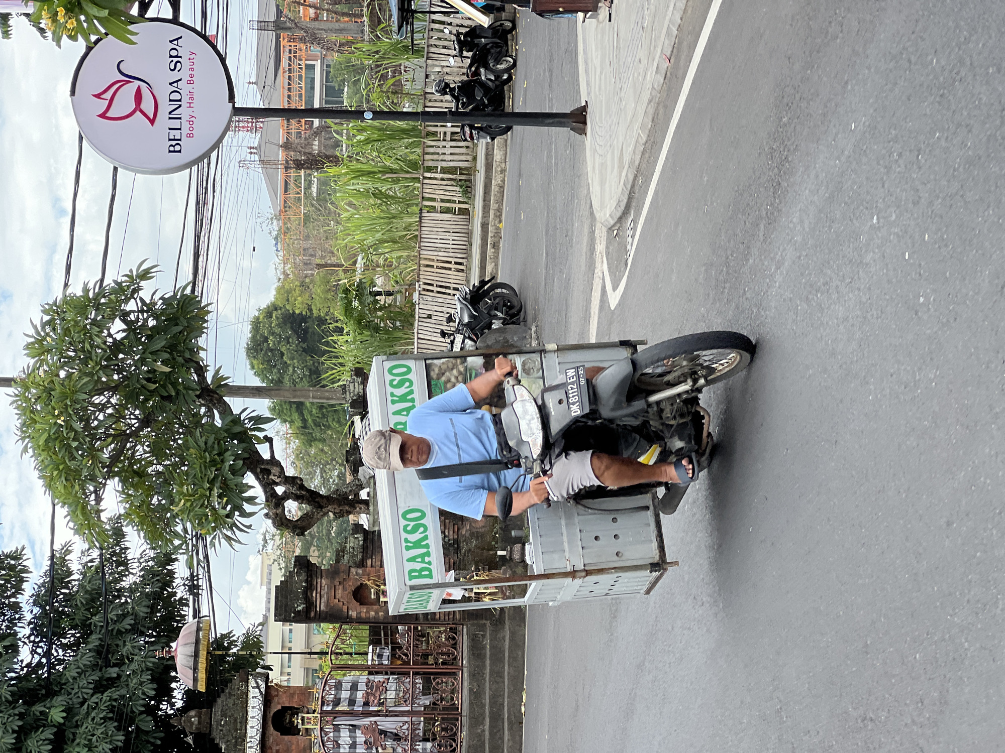 A man rides a motorcycle with a food cart attached, on a street in Denpasar, Indonesia. The man, wearing a blue t-shirt, gray shorts, a baseball cap, and sandals, is driving the motorcycle and appears to be operating the food cart. The cart is labeled "BAKSO" in green lettering on a white background. There are several motorcycles parked on the side of the road, and a sign for "BELINDA SPA" is visible in the background. The street is paved, with white lane markings and a curb. The environment appears to be an urban or semi-urban area with trees, buildings, and a cloudy sky.