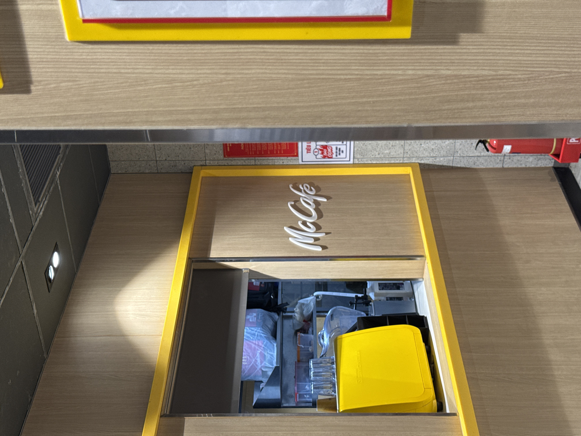This is a medium shot of the McCafe counter at a McDonald's in Nha Trang, Vietnam. The counter is made of light-colored wood with a bright yellow trim. On the left side, a yellow coffee machine is visible, with a stack of clear plastic cups on top. Behind the counter, shelves are stocked with various items, including plastic bags, containers, and bottles. The "McCafé" logo is displayed in white script on the wooden wall. To the right of the counter, a red fire extinguisher is visible, along with a sign that reads "TIỂU LUẬN" and "BẢO ĐỘNG" with a red warning icon. The ceiling has a grid of dark panels with two spotlights.