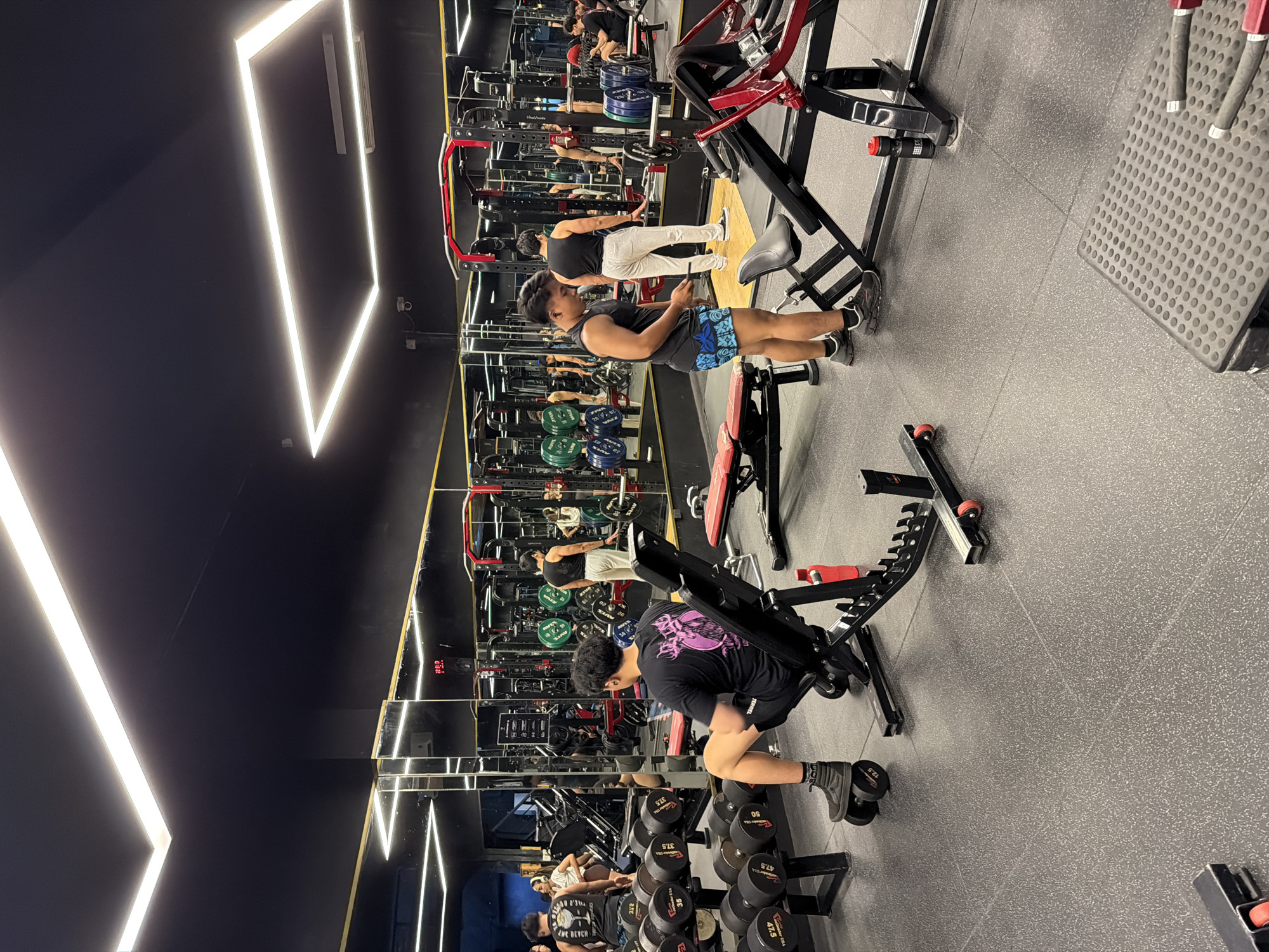 Here's a detailed description of the image:
**Setting:** The image appears to be taken inside a modern gym, possibly in Denpasar, Indonesia. The gym has a sleek aesthetic with black walls and distinctive, angular LED lighting fixtures on the ceiling. Large mirrors line one side of the room. The floor is a speckled grey, likely a rubberized material suitable for gym use.
**People:** There are several people visible in the image, mostly men. Their actions seem to be focused on exercising.
* A man is seated on a weight bench, potentially lifting dumbbells. He is wearing a black t-shirt and shorts.
* Another man stands near a weight machine, focused on his phone. He is wearing a tank top and patterned shorts.
* Other individuals are further in the background, reflected in the mirrors, and they also seem to be working out.
**Objects:**
* **Weight Machines:** There are various weight machines in the gym, including a leg press machine, a seated row machine, and a smith machine.
* **Dumbbells:** Racks of dumbbells of varying weights are visible near the foreground. Weights are stacked in an organized way.
* **Weight Bench:** An adjustable weight bench is positioned in the foreground.
* **Mirrors:** Large mirrors line the wall, reflecting the gym and the people inside.
**Actions and Interactions:** The primary action is individuals working out in the gym. The gym is populated but not overly crowded.
