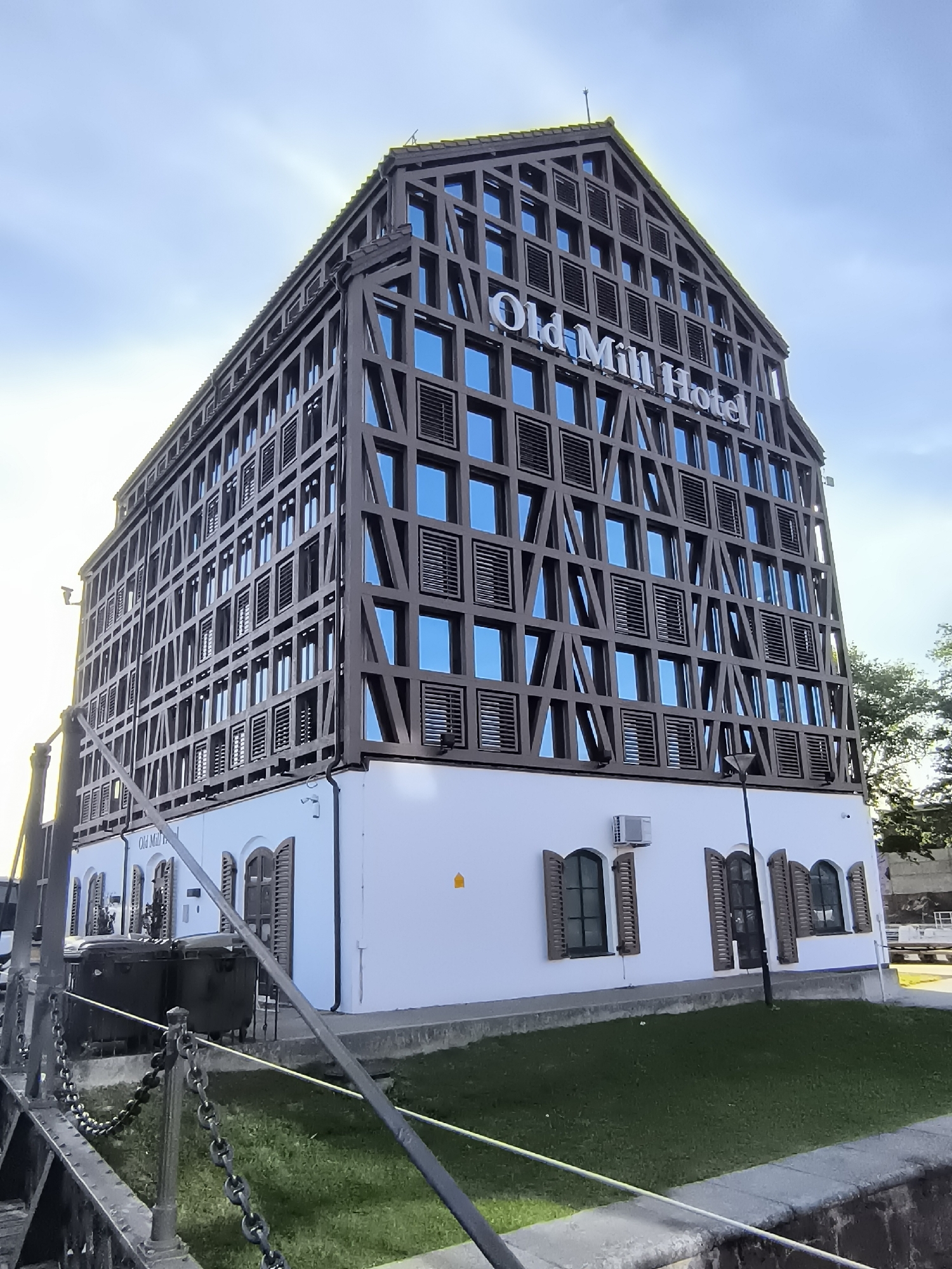 The image shows the Old Mill Hotel in Klaipėda, Lithuania, an architecturally unique building with a modern, geometric facade overlaid on a traditional timber-frame design. The hotel is a tall, multi-story structure with a prominent gabled roof. The upper floors feature a dark wooden lattice pattern encasing numerous windows with blue glass and some with louvered shutters. The name "Old Mill Hotel" is displayed in large white lettering on the upper section of the building. The lower part of the building is a white, smooth-walled base with arched windows fitted with brown wooden shutters. In front of the building, there is a strip of green grass bordered by a concrete edge. To the left of the hotel, a metal railing with a thick chain runs along the foreground, suggesting proximity to a waterway or walkway. Two dark, industrial-looking bins are visible behind the railing. The sky is bright blue with some wispy clouds, and the sun is shining, casting a bright glare on the left side of the hotel. Trees can be seen to the right of the building. The scene appears to be a daytime exterior shot in an urban setting.