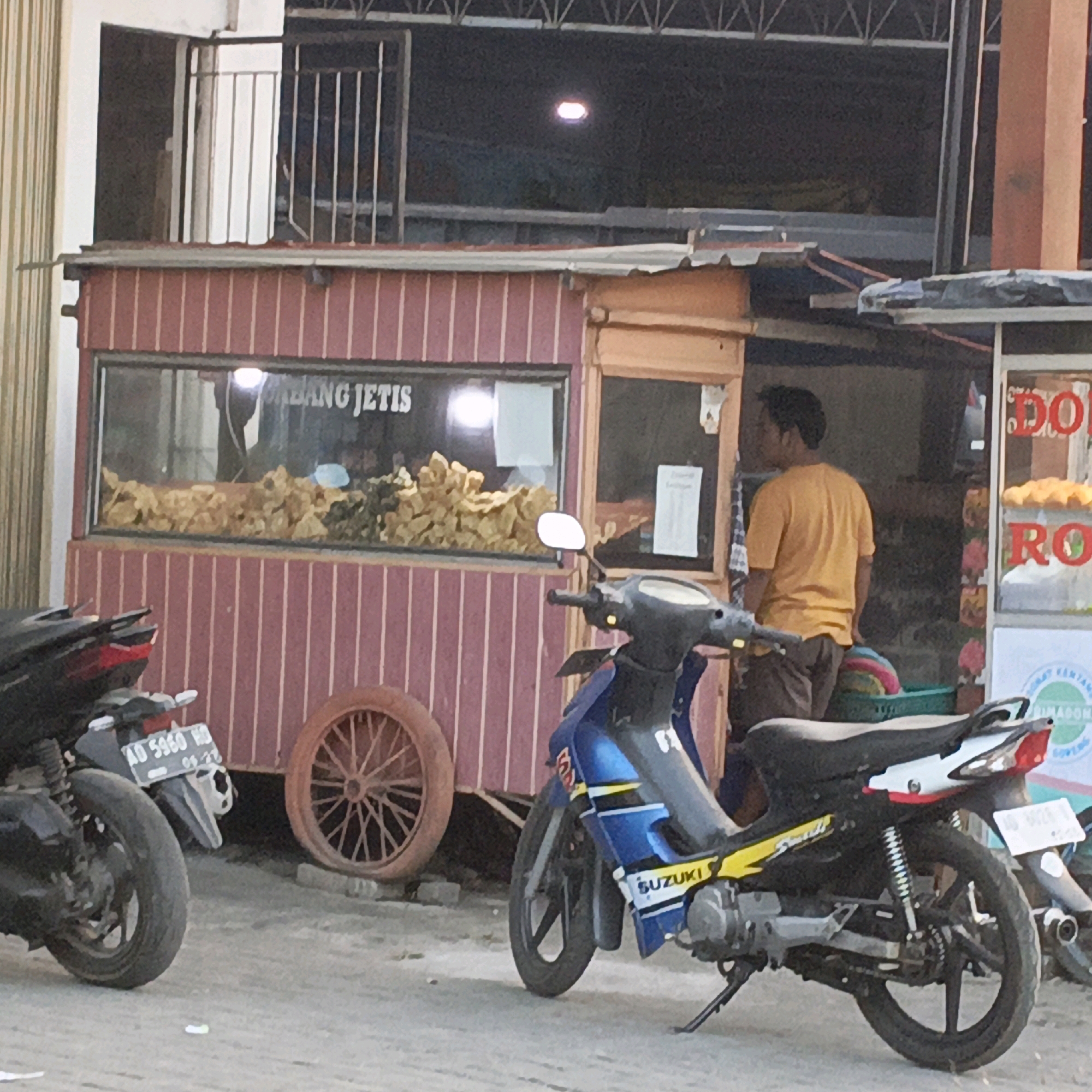 In Gedangan, Indonesia, a street vendor stall selling food is depicted. The stall has a red and white striped exterior and a glass display case filled with food items. A man in a yellow shirt stands behind the counter, facing away from the camera. Two motorcycles are parked in front of the stall. The motorcycle on the left is black and has a license plate that reads "AD 5960 WD". The motorcycle on the right is blue and white and has the brand name "SUZUKI" visible on its side. In the background, a large, rusty wheel is leaning against the stall. The scene appears to be during the daytime, with artificial lighting from inside the stall illuminating the food.