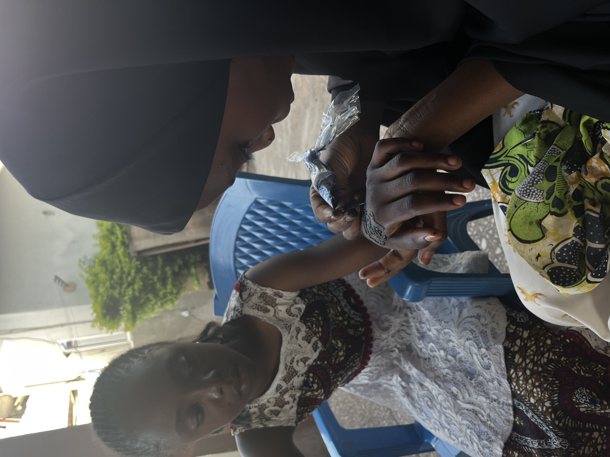 In Jos, Nigeria, a woman wearing a black hijab and black clothing is applying henna to the hand of another person. The person receiving the henna is seated on a blue plastic chair and wearing a patterned dress with white lace details. The henna is being applied from a plastic cone. In the background, a building and green foliage are visible.