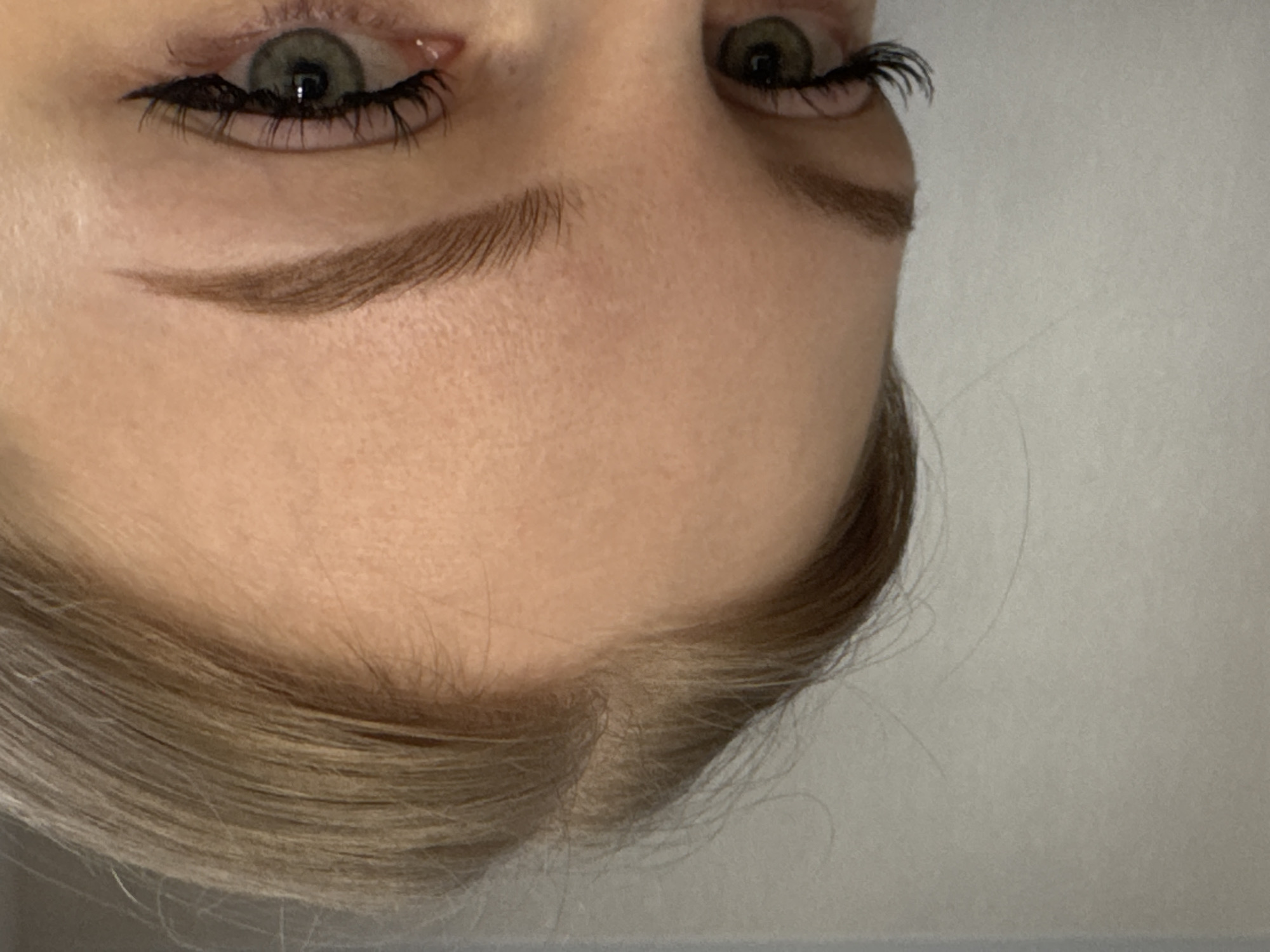 A close-up, inverted photograph depicts the upper portion of a person's face. The subject has light skin, green eyes with black eyeliner and false eyelashes, and light brown eyebrows. The upper part of their blonde hair is visible near the bottom of the image. No other details are discernible to identify a specific location like Perm, Russia.