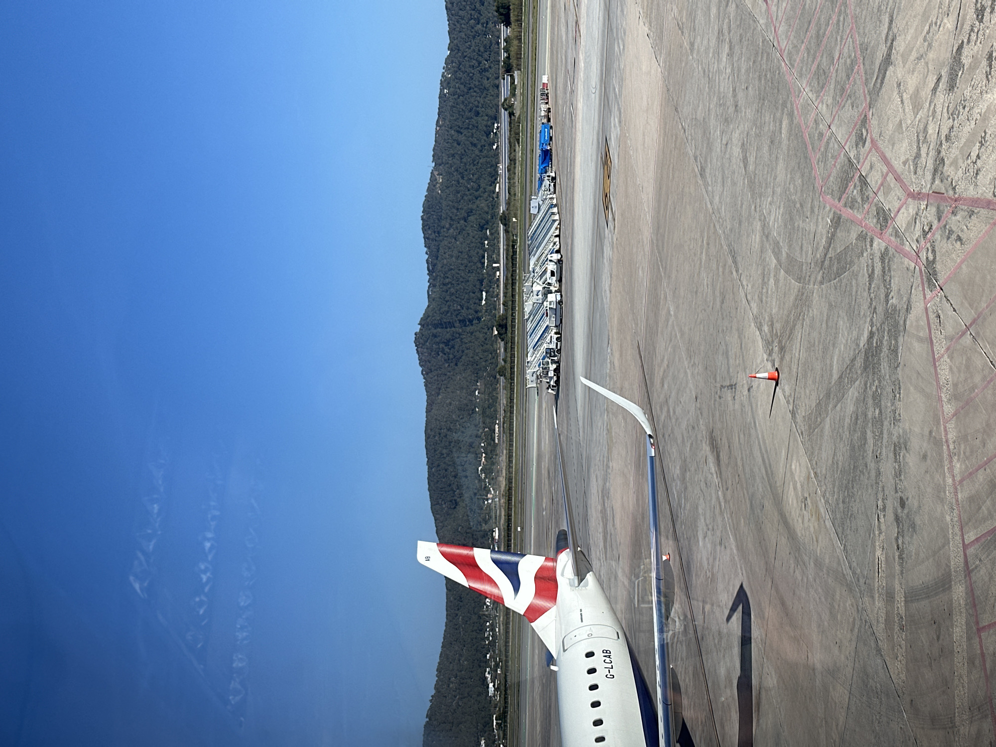 A British Airways aircraft, tail number G-LCAB, is positioned on the tarmac, likely at Ibiza Airport in Sant Josep de sa Talaia, Spain. The aircraft is viewed through a window. The plane's tail, adorned with the Union Jack, is visible. Part of the wing with the winglet is also visible. Beyond the tarmac, a series of airport support vehicles are parked near a building. A treed mountain is behind the airport facilities. The sky is blue. An orange and white traffic cone is sitting on the tarmac near the plane. Paint markings are visible on the tarmac.