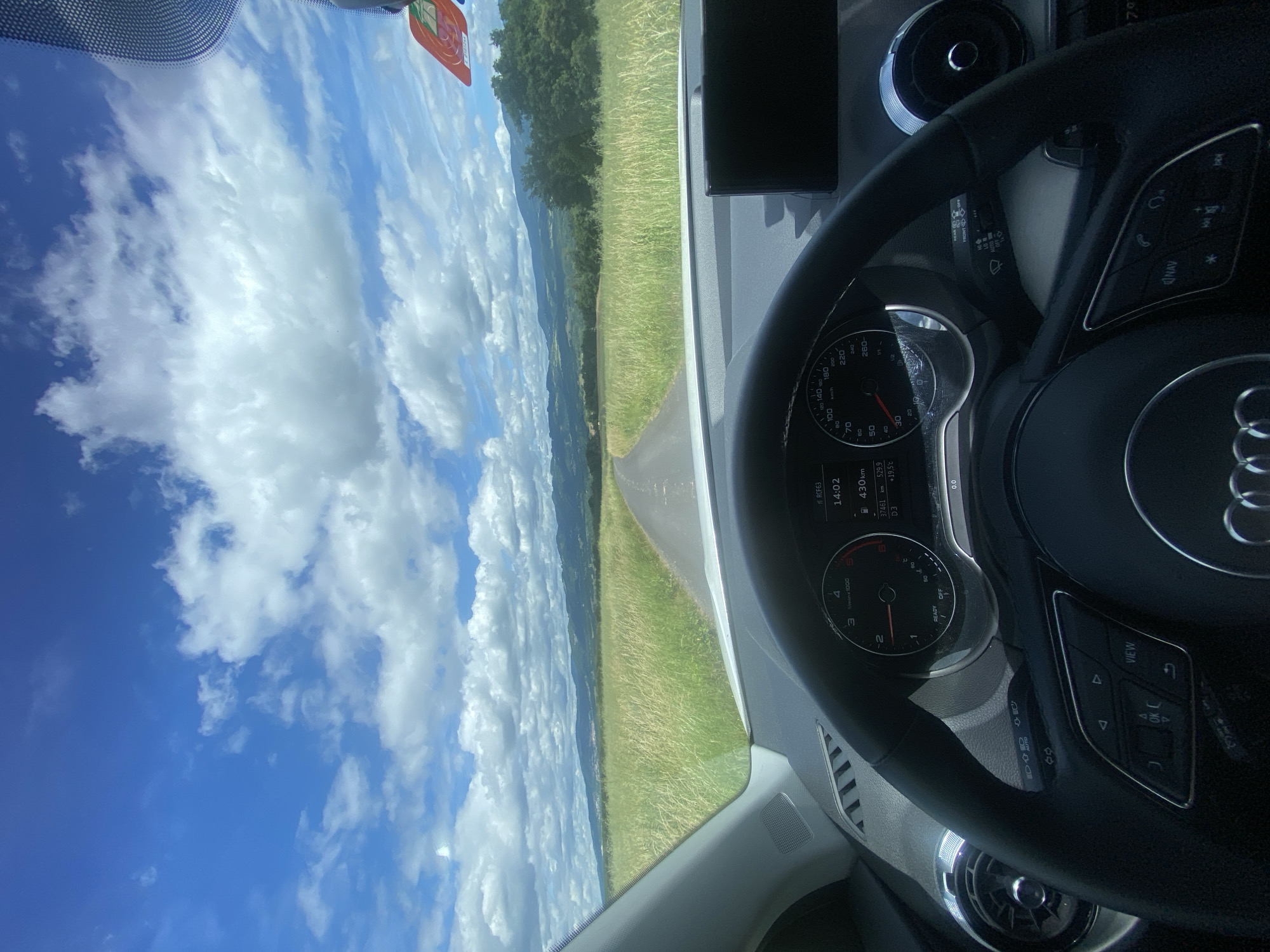 The image provides a first-person perspective from inside a car, looking out through the windshield onto a scenic road and landscape. The right side of the frame is dominated by the car's interior, featuring an Audi steering wheel and a partial view of the dashboard.
On the dashboard, the time is clearly visible as "14:02," indicating early afternoon. Other readings include a total odometer of "37461 km," a trip meter showing "529.9 km," and an outside temperature of "+19.5°C." The speedometer, marked in "km/h," and a tachometer displaying "READY" are also visible. The steering wheel features control buttons with labels like "NAV," "VIEW," and "OK," along with icons for phone, volume, and track control. A wiper/indicator stalk is visible behind the steering wheel, showing settings such as "OFF" and "AUTO."
Beyond the car, a bright blue sky with numerous large, fluffy white clouds fills the upper portion of the exterior view. A narrow, paved road stretches forward through a predominantly green landscape. Fields of tall grass, varying in shades of green and golden, line both sides of the road. In the distance, rolling hills and mountains rise against the horizon, partially veiled by the clouds.
The scene depicts a calm and pleasant drive during the daytime, under sunny and clear weather conditions. The vehicle is evidently moving through a rural or semi-mountainous region. The use of kilometers per hour and the natural landscape are consistent with the provided location context of Courpière, France. The overall impression is one of serene travel through a picturesque environment.