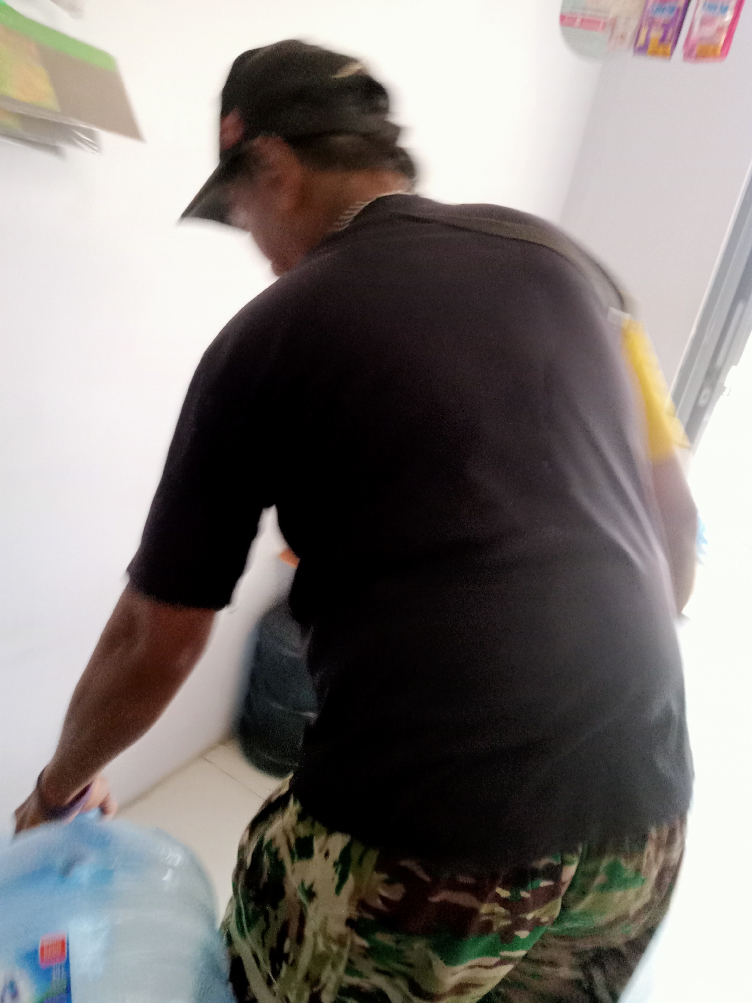 A person wearing a black t-shirt, a black baseball cap, and camouflage pants is seen from behind, holding a large blue water jug. The person appears to be in motion, as indicated by the motion blur in the image. The background consists of a white wall and a light-colored floor. In the background, there are also some shelves with items on them. The overall setting appears to be indoors, possibly a home or a store. The image quality is poor due to motion blur and low resolution, making it difficult to discern specific details about the person's face or the surrounding environment. The location in Simo, Indonesia cannot be determined from the image alone.
