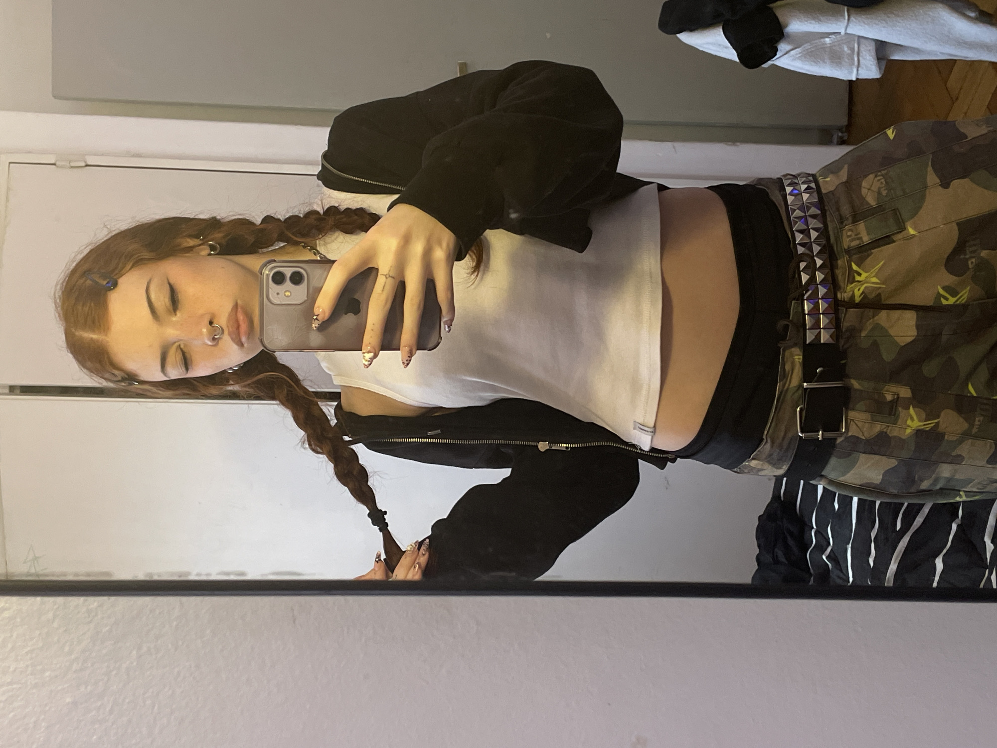 A young woman is reflected in a mirror. She has long, braided auburn hair and is wearing a white tank top, a black zip-up jacket, and camouflage pants with a studded belt. She is holding a smartphone in her right hand and is taking a selfie. Her left hand is at the end of her left braid. She has a nose ring. The background includes a white door and wall, and some clothing on a surface behind her. There is no evidence to support the location is Buenos Aires, Argentina.