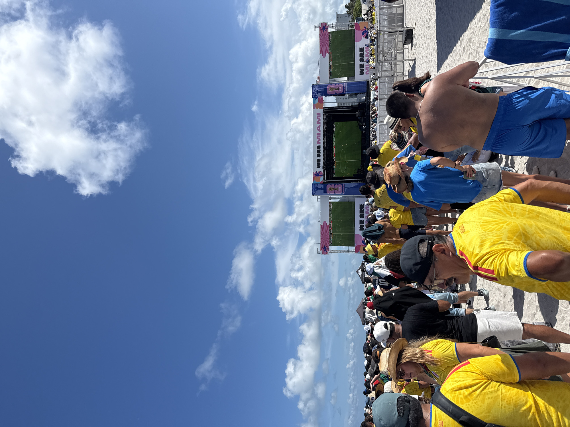 An outdoor crowd watches a soccer match on large screens set up on Miami Beach. The screens are part of a "WE ARE MIAMI" event. People in the foreground are wearing yellow shirts, and some are wearing hats. The beach is sandy, and the sky is blue with scattered white clouds. In the background, there are buildings and palm trees. The event appears to be a public viewing of a sports game.