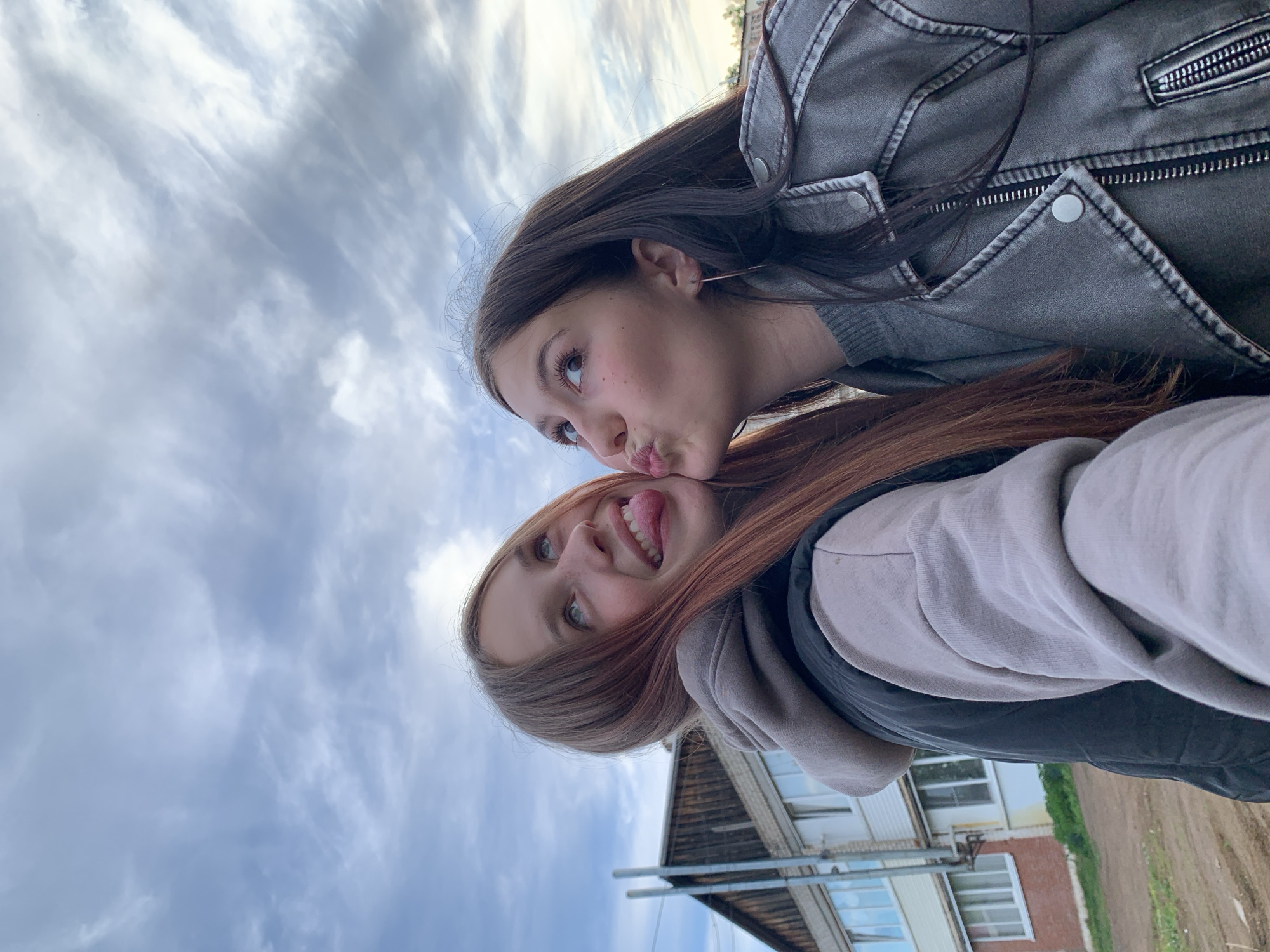 Two young women are posing for a selfie. One woman has brown hair and is wearing a gray jacket. She is making a kissing face. The other woman has auburn hair and is wearing a gray hoodie. She is sticking her tongue out. In the background is a cloudy sky and the edge of a building. The location is likely Фоки, Russia.