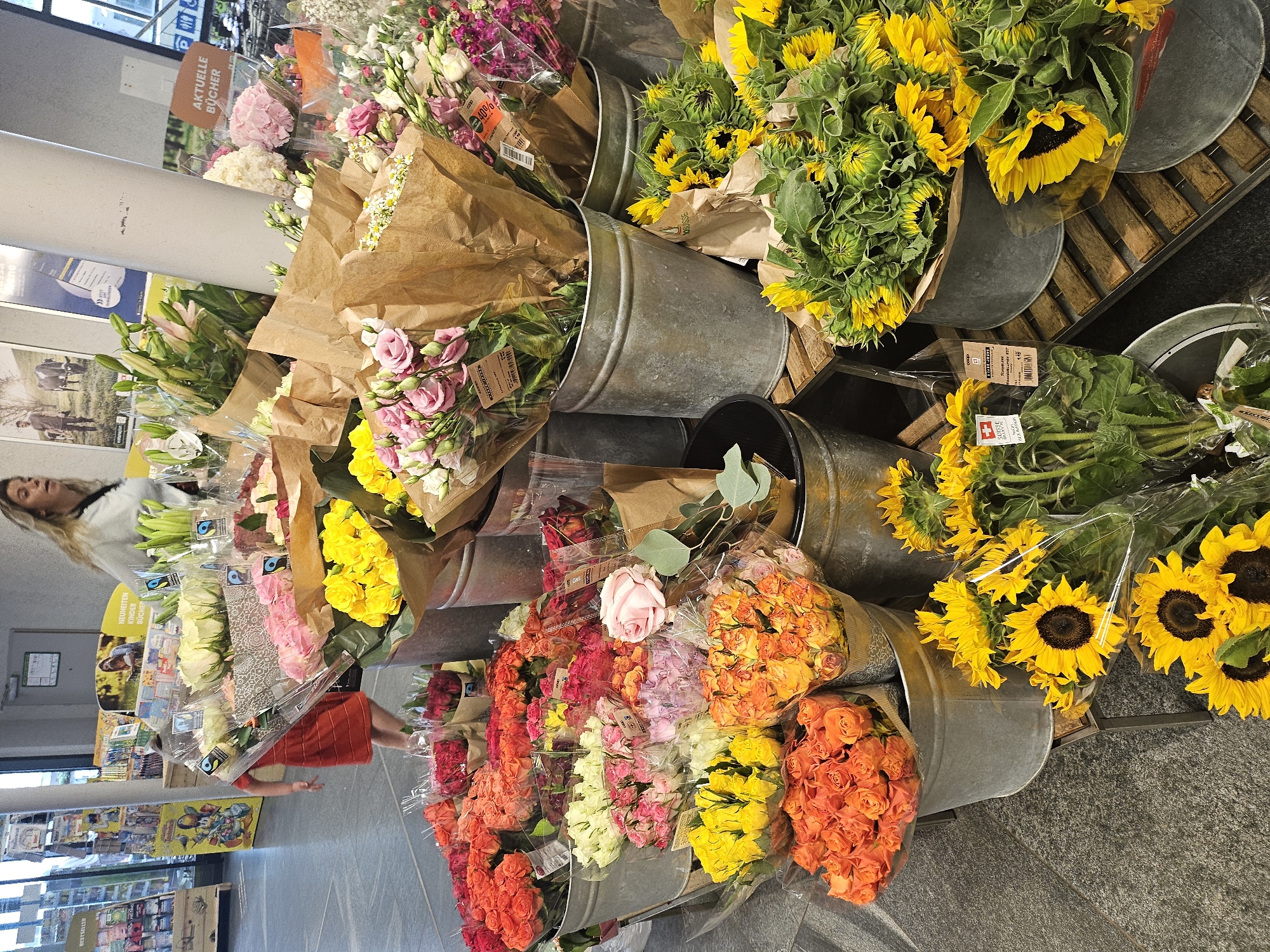 The image shows a flower stand in what appears to be a supermarket or outdoor market, with a woman in the foreground. She has blonde hair and is wearing a white top, looking to her left as she walks past the display. The flower stand is abundant with various bouquets of fresh flowers, arranged in galvanized metal buckets and brown paper wrapping.
Prominently featured are vibrant sunflowers with bright yellow petals and dark centers, as well as bouquets of pink and yellow roses, and other colorful blooms like hydrangeas and lilies. Some flowers are packaged in clear plastic. Several buckets of flowers are placed on wooden planks, creating a tiered display.
In the background, shelves stocked with products are visible, suggesting a retail environment. One prominent sign in German reads "AKTUELLE BÜCHER" which translates to "CURRENT BOOKS". Other signs and labels are present, including one with a Swiss flag symbol and text, likely indicating the origin or branding of the flowers. The overall scene suggests a bright, well-lit environment, possibly during daytime hours. The presence of fresh flowers and the organized display imply a retail setting focused on selling flowers.