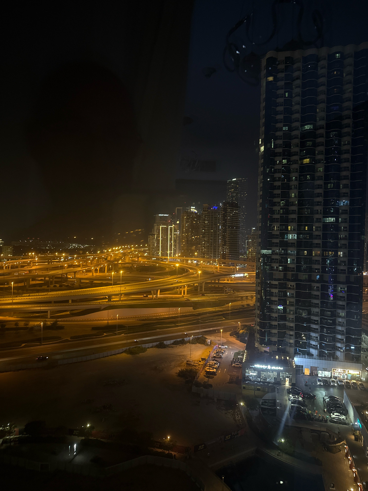 The image captures a nighttime view of a complex highway interchange in Dubai, United Arab Emirates, with a tall, modern apartment building dominating the foreground on the right. The highway, illuminated by numerous streetlights, features multiple elevated lanes and curves, suggesting a busy urban transportation hub. The apartment building is characterized by its dark facade with repeating balconies and numerous illuminated windows, indicating residential occupancy. In the background, other illuminated high-rise buildings of Dubai's skyline are visible, contributing to the city's characteristic dense urban landscape. The overall scene is a testament to Dubai's modern infrastructure and urban development.