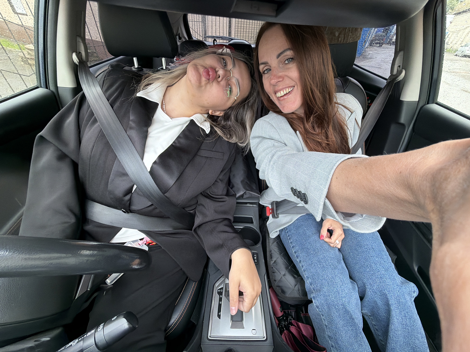 Two women are featured in a selfie taken from inside a car, likely in Birobidzhan, Russia. The woman on the right, who appears to be taking the photo, smiles broadly at the camera. She has long brown hair and is dressed in a light grey blazer over a lighter top and blue jeans. Her right arm, holding the camera, is visible in the foreground. Beside her, the woman on the left, with blonde-streaked hair and glasses, leans her head and playfully pouts towards the lens. She wears a black blazer over a white collared shirt. Both women are buckled into their seatbelts.
The interior of the car is visible, showing dark grey seats, headrests, a central console with an automatic gear shifter (displaying "P R N D-M"), and an armrest. A maroon umbrella is tucked near the passenger's feet. The car windows show numerous water droplets, indicating rainy or recently rainy weather outside. The blurred exterior view includes a fence, some greenery, and indistinct structures under what appears to be an overcast sky. A control stalk near the driver's position shows partial text like "AUTO" and "OFF". The expressions of the women suggest a lighthearted and cheerful mood.