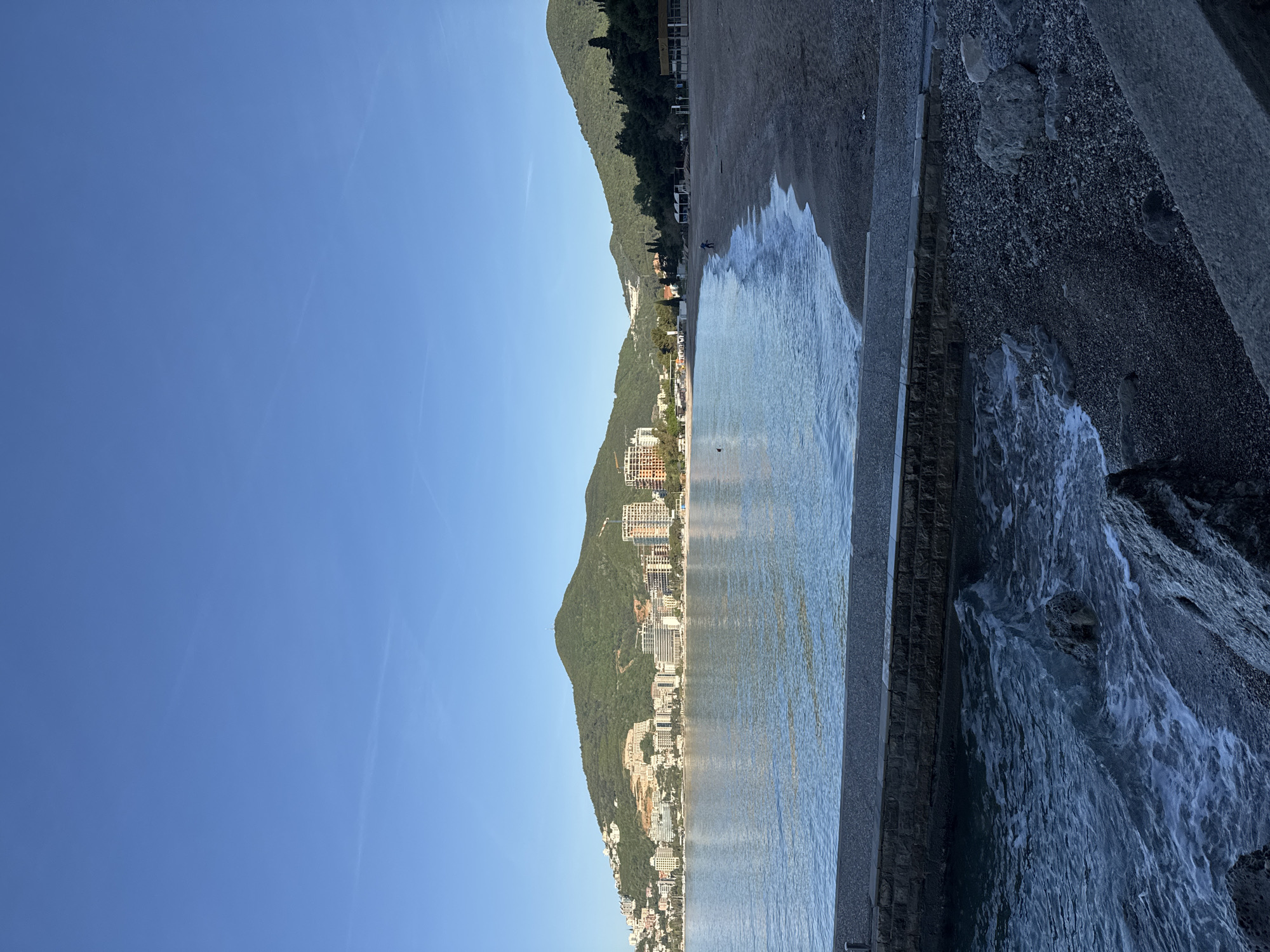 The image shows a coastline in Budva, Montenegro, on a sunny day. The clear blue sky occupies the left portion of the image. A mountain range runs along the upper part of the image, covered in green vegetation. Below the mountains, there is a series of buildings and hotels stretching along the coastline. The sea is visible on the right side of the image, with small waves lapping against a rocky shoreline and a gray stone wall. The photo is rotated 90 degrees.