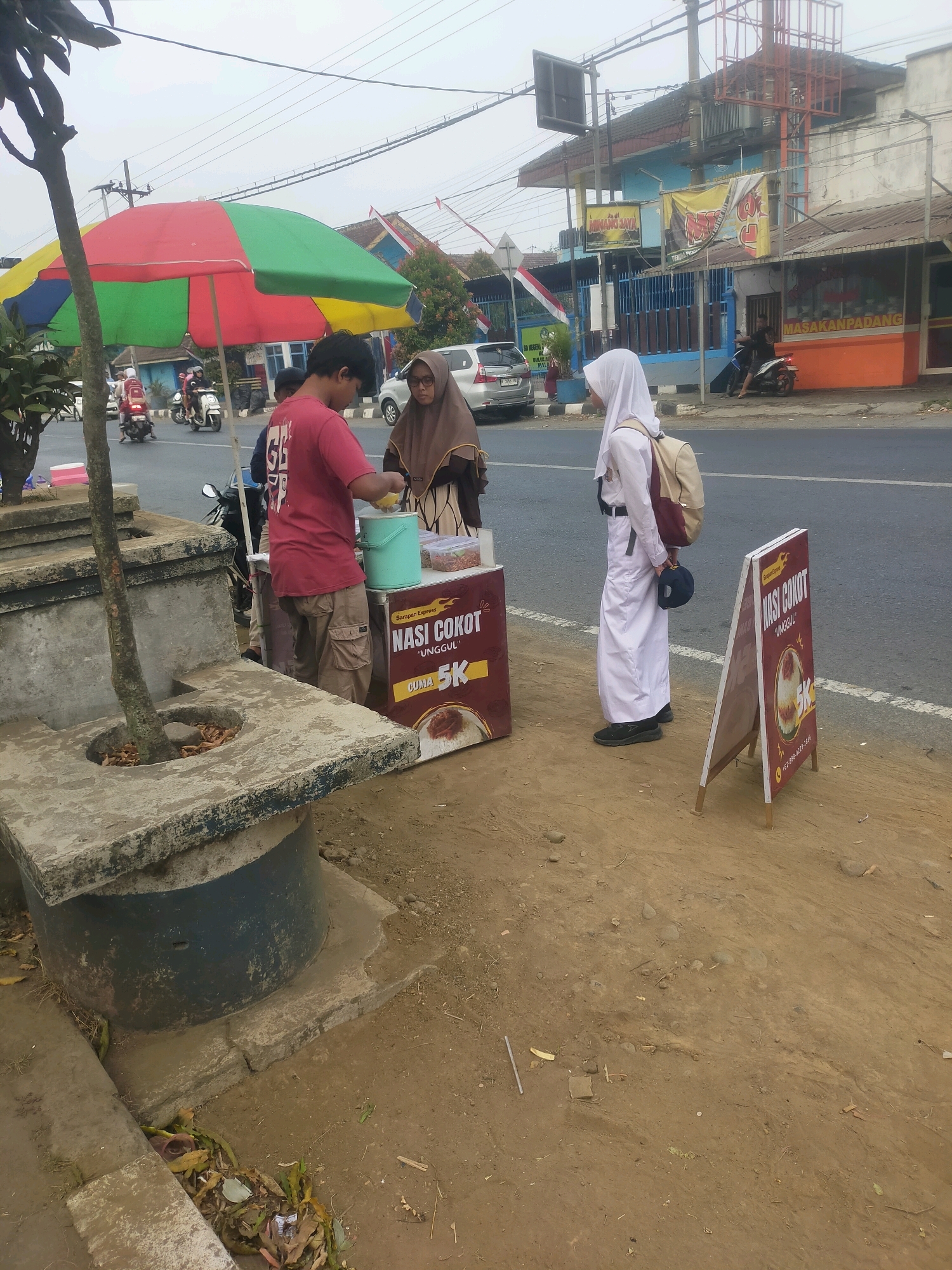 The image depicts a street scene in Bululawang, Indonesia, where a food vendor is operating. In the foreground, a young person in a red t-shirt and cargo pants is serving food from a mobile cart labeled "Nasi Cokot Unggul". They are interacting with a woman wearing a hijab and a white uniform, who appears to be a student. The cart is decorated with images and text advertising the food, including the price "5K". A sign in the shape of an A-frame, also advertising "Nasi Cokot", is placed on the side of the road. Behind the vendor and customer, a colorful umbrella provides shade. In the background, there is a road with passing motorcycles and cars, and buildings along the side of the street. One building has signs for "MASAKANPADANG" and "MINANG JAYA". The overall environment suggests a busy street with commercial activity.