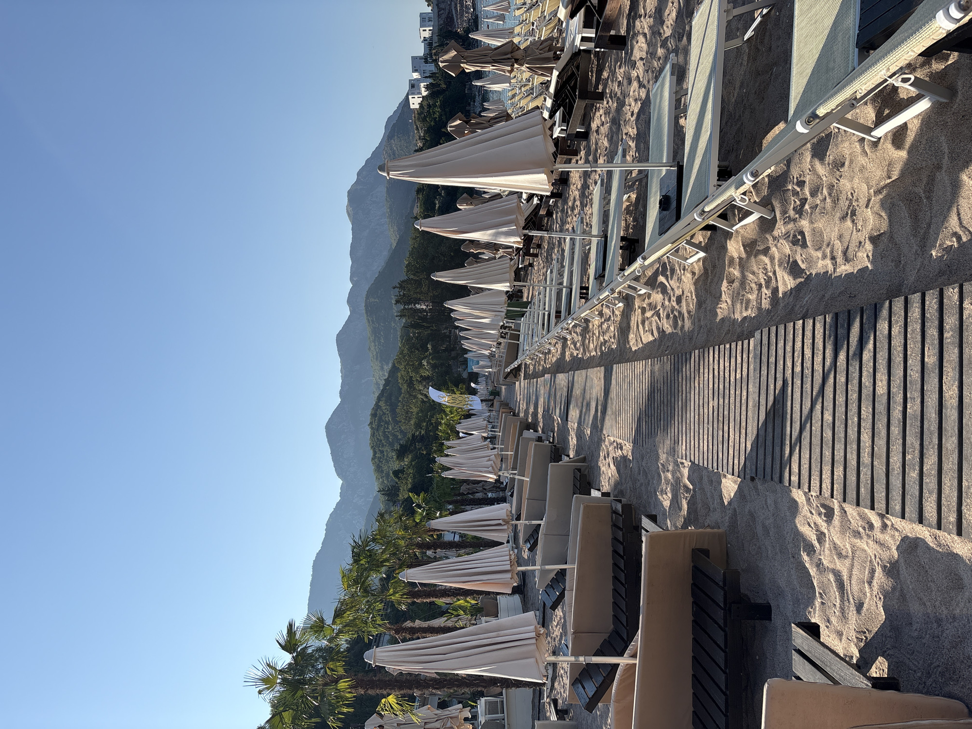 This is a photograph of a beach in Kamenovo, Montenegro, taken from a low angle, looking down a wooden boardwalk. The boardwalk is lined on both sides with rows of closed beach umbrellas and lounge chairs, which are predominantly white and beige. Palm trees are visible to the left of the boardwalk, and in the background, there are green, forested mountains with some buildings on the hillsides. The sky is a clear, bright blue. The sand is light brown and appears undisturbed. The scene is set during the daytime, with sunlight casting distinct shadows from the boardwalk onto the sand. There are no people visible in the image.