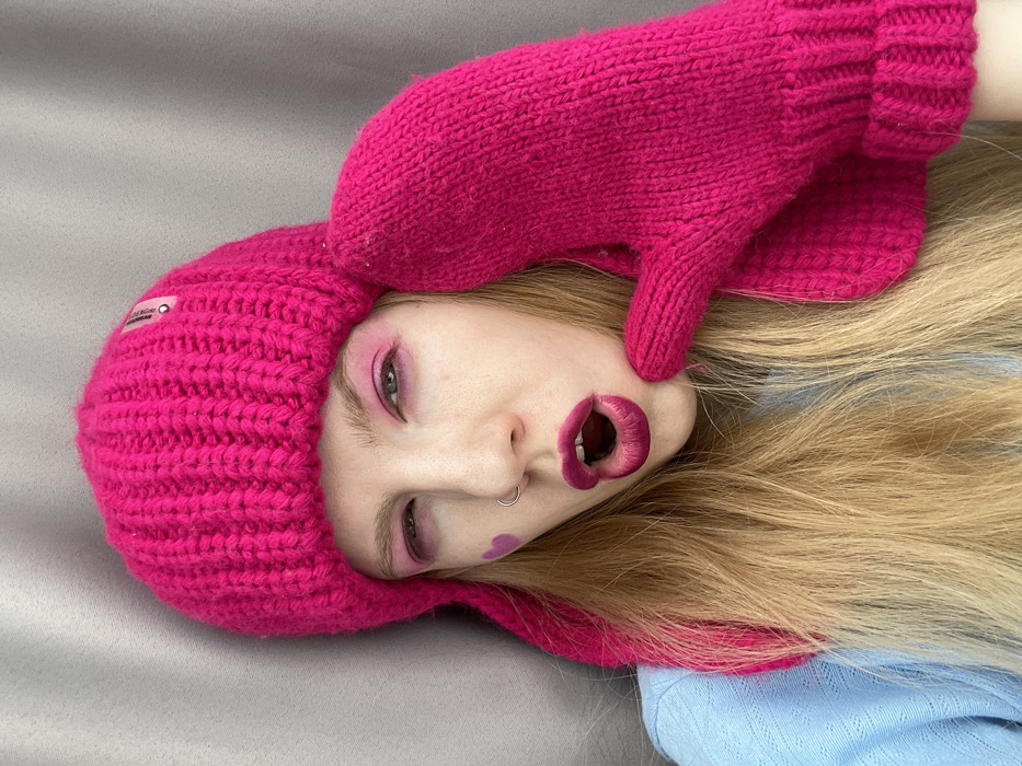 An individual with long blonde hair, appearing to be a young adult female, is depicted in a close-up, head-and-shoulders portrait. She wears a fuchsia pink, ribbed knitted beanie hat with ear flaps, which features a small, translucent rectangular tag on the upper right side. A matching fuchsia pink knitted mitten covers the right side of her face and neck, with the hand positioned as if touching her cheek. The individual is dressed in a light blue long-sleeved garment with a subtle patterned texture.
Her facial features include prominent fuchsia pink eyeshadow applied to both eyelids, with some appearing smudged or intentionally extended. Her lips are coated in a dark fuchsia lipstick and are open, forming an 'O' shape, revealing the upper and lower teeth and a portion of the tongue. A silver nose ring is present in her left nostril. A small, solid purple heart shape is drawn on her left cheekbone, below the eye. Her eyes are partially closed or squinted.
The background consists of a plain, solid gray surface, possibly a wall or fabric, providing a stark contrast to the subject's vibrant attire and makeup. Illumination is even, suggesting an indoor setting. The precise location within Izhevsk, Russia, is not discernible from the visual information presented in the image.