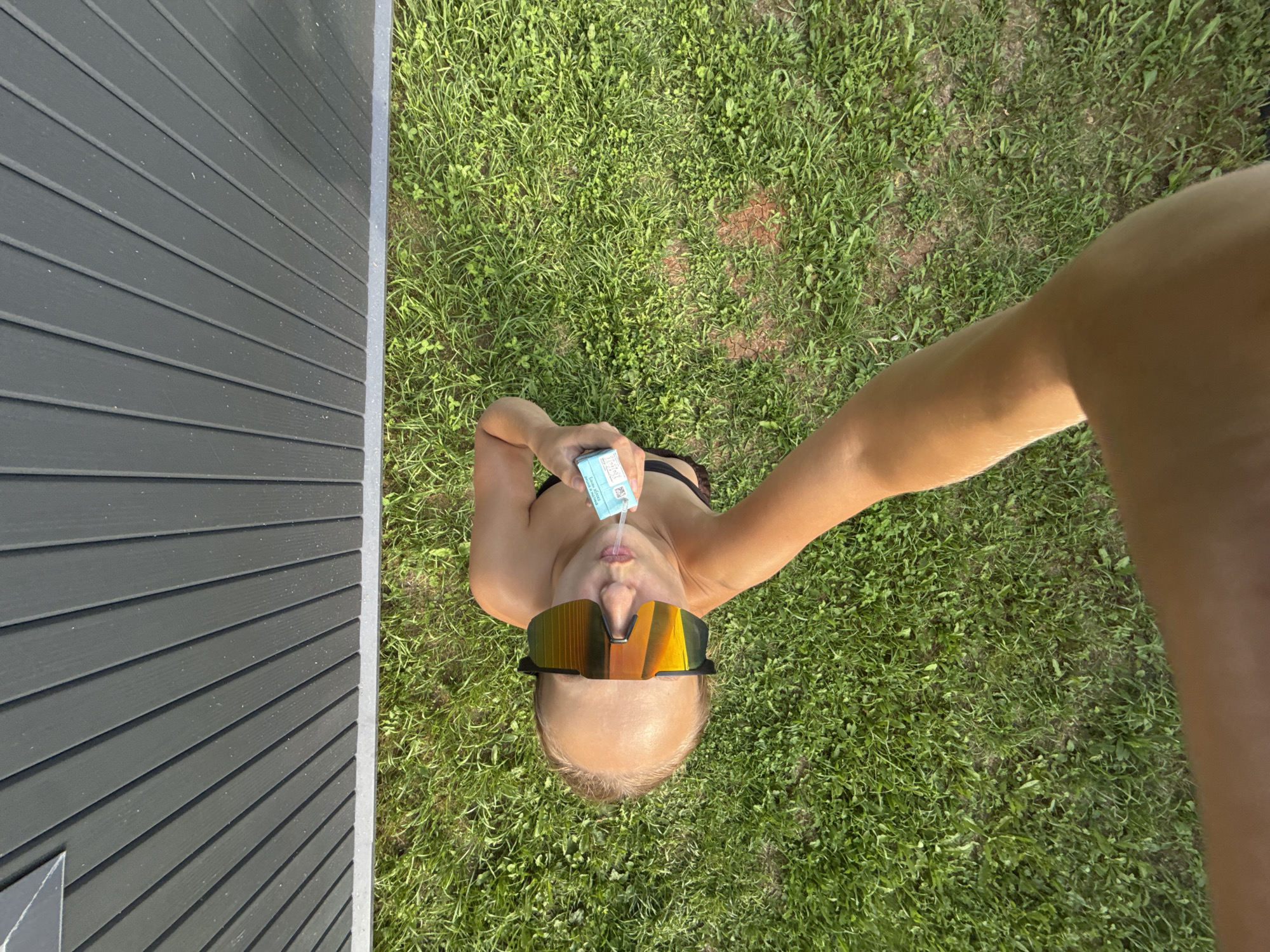 In the city of Podshivalovo, Russia, a person with a bald head and wearing large, reflective sunglasses is lying on a patch of green grass. They are holding a small, light blue box with white text and a straw to their mouth, possibly administering a liquid. The person is also wearing dark-colored swimwear. Behind them, a dark gray, slatted wall of a building is visible, with the grass extending towards it. The image is taken from a high angle, looking down at the scene.