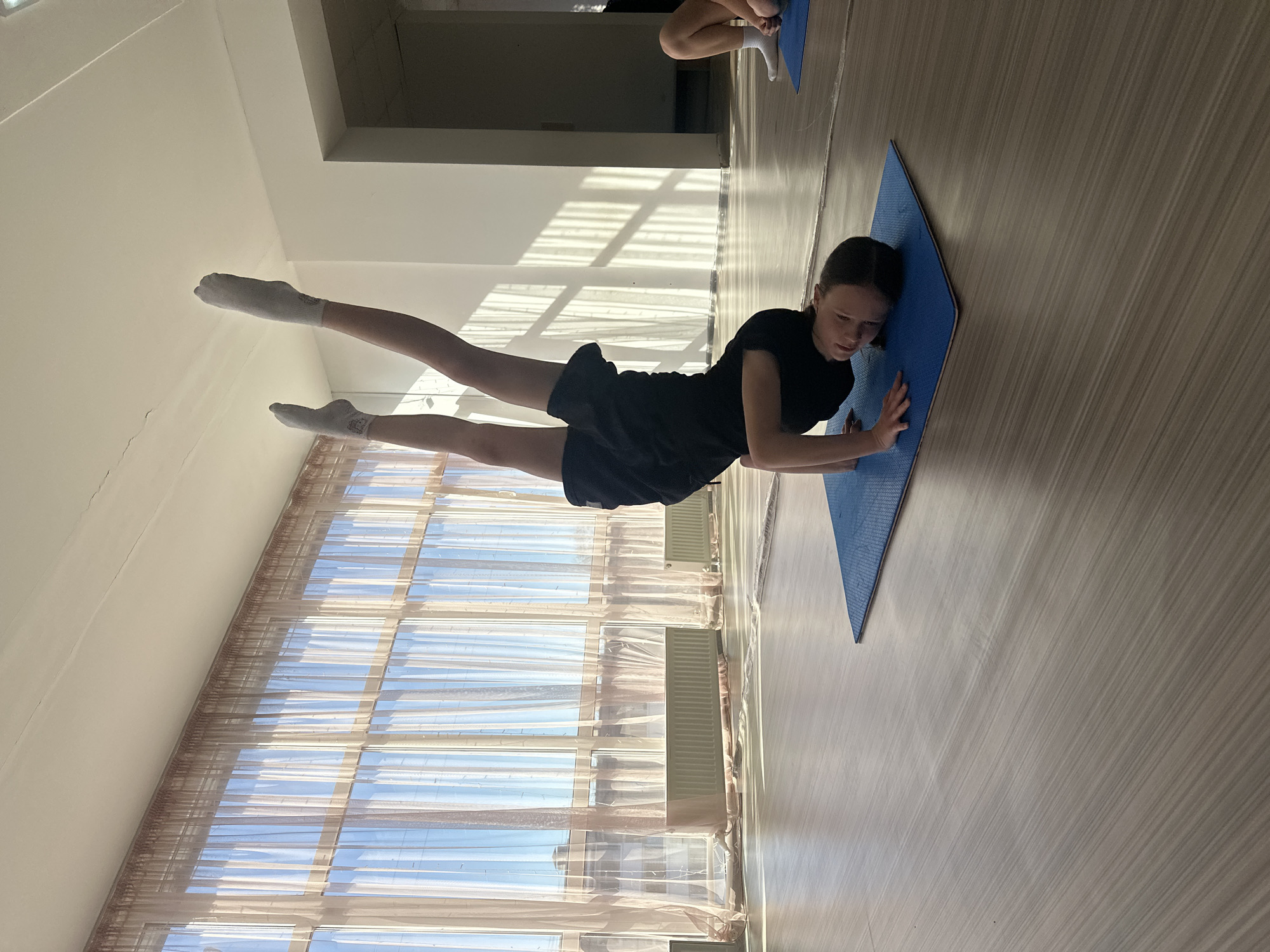 A young girl in a black shirt and black shorts is doing yoga or stretching on a blue yoga mat inside a room. She is lying on her stomach with her legs raised and feet pointed upwards. She is wearing white socks. In the background, a window with sheer curtains lets in sunlight, creating shadows on the wall. The location is Votkinsk, Russia. Another person is partially visible in the background, also on a yoga mat. The floor is light-colored.