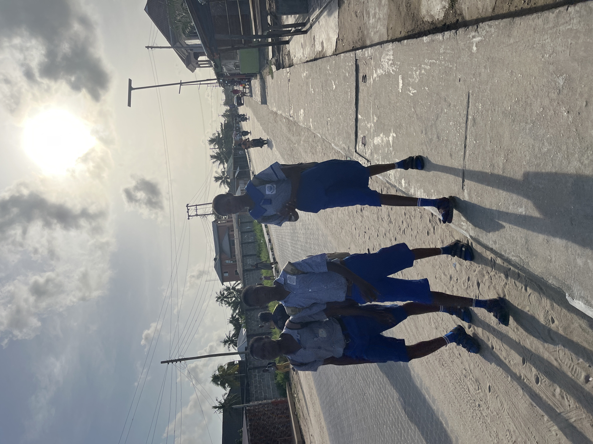 Three boys in blue and white checkered school uniforms are walking along a concrete sidewalk in Badagry, Nigeria. They are walking towards the right side of the frame, away from the camera. The background shows buildings, palm trees, utility poles, and a bright sky with clouds. The sidewalk is cracked and worn, with visible dirt and sand. The sun is bright, casting strong shadows of the boys on the ground.