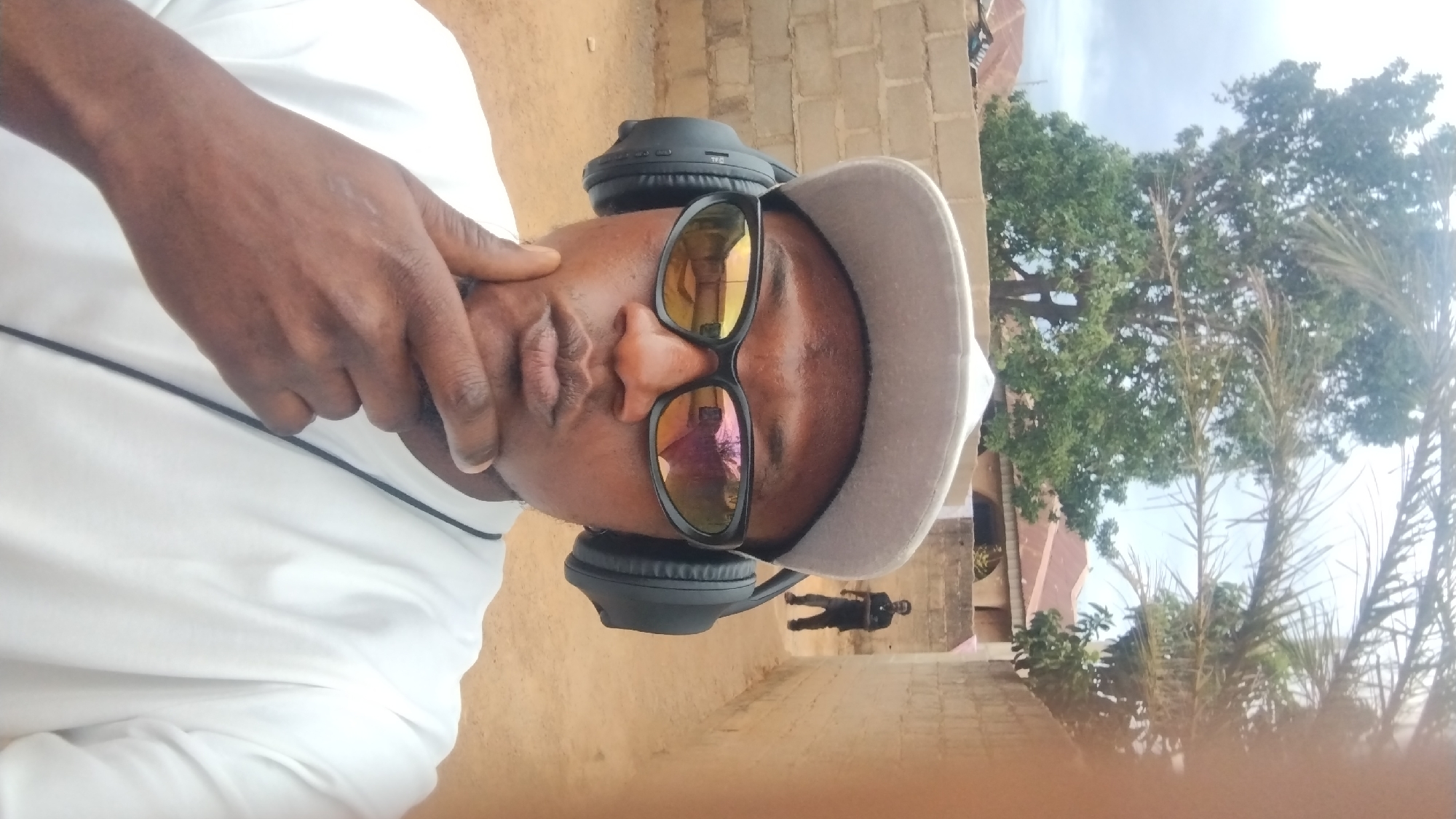 A man wearing sunglasses, a cap, and headphones poses for a selfie outdoors. The reflection in his sunglasses shows a building with a red roof. Behind him, a blurry figure walks along a path beside a brick wall. Trees and foliage form a backdrop. The image was likely taken in Jos, Nigeria.