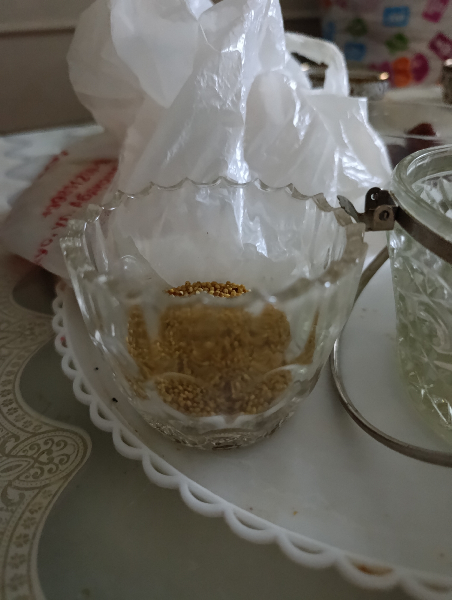 A close-up, top-down shot captures a small, clear glass bowl filled with small, light-brown seeds. The bowl has a decorative, scalloped edge and sits on a white, scalloped-edged tray. To the right of the bowl, a portion of a larger, clear glass container with a textured surface is visible, held by a metal clamp attached to the bowl's edge. In the background, a crinkled white plastic bag is prominent, partially obscuring a patterned surface beneath it. The scene appears to be indoors, possibly on a table. There are no people visible in the image. The overall lighting is bright, with some areas of shadow. The specific location in Nukus, Uzbekistan, cannot be determined from the visual information provided.