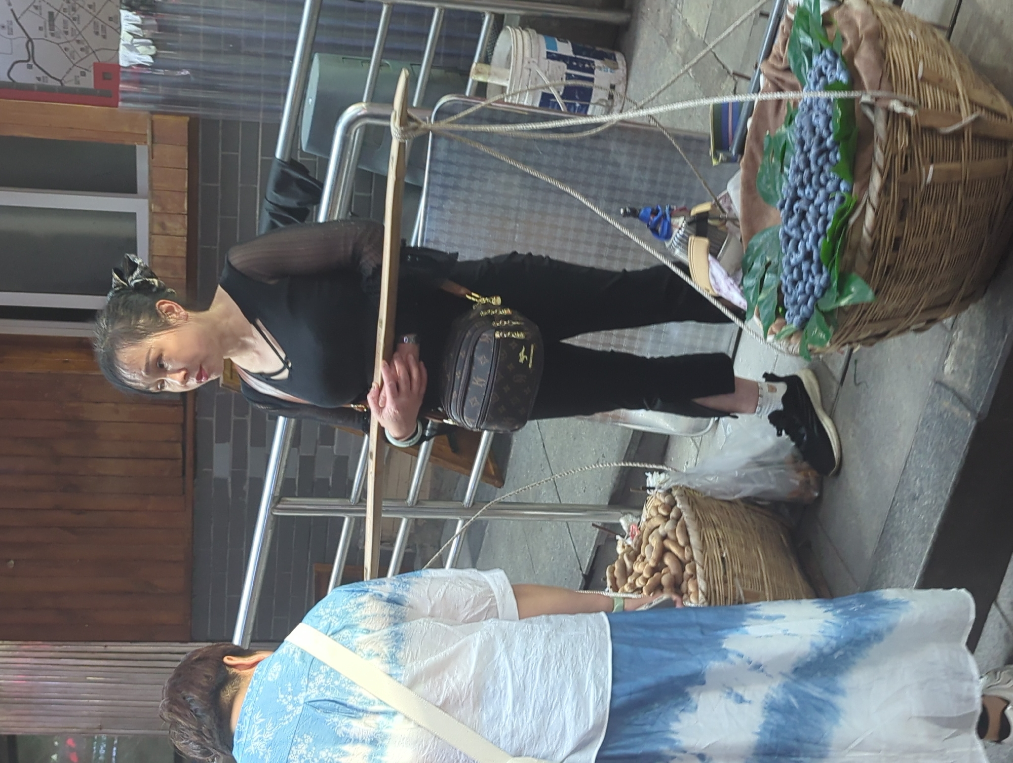This vertical image captures a street scene, likely a market or public walkway, in Yuzhong District, China. The primary subjects are two women.
The woman in the foreground, facing left, wears a black top with sheer sleeves and black pants. She has her hair tied back with a patterned scrunchie and appears to be wearing a jade-colored bracelet on her left wrist. She holds a wooden carrying pole that extends across the frame. From the pole hang two large, woven baskets by ropes. One basket prominently displays artificial bunches of blue/purple grapes nestled among green leaves. The other basket, partially visible, contains a mound of light brown, elongated, tuber-like items, possibly peanuts or taro. She carries a brown patterned shoulder bag with "LV"-style monograms. She is wearing white socks and black and white sneakers.
Standing opposite her, mostly seen from behind, is another person wearing a white and blue tie-dye top or dress. Their hair is dark and short. This person is looking down, possibly at the goods.
The background features various urban elements: metal railings, a grey brick wall, wooden paneling, and a white window. A white bucket with blue markings is visible behind the railings. On a wall in the upper left, a blurry map with street patterns and some unreadable text is mounted. The ground is paved with stone or concrete. The scene is brightly lit, indicating daytime.