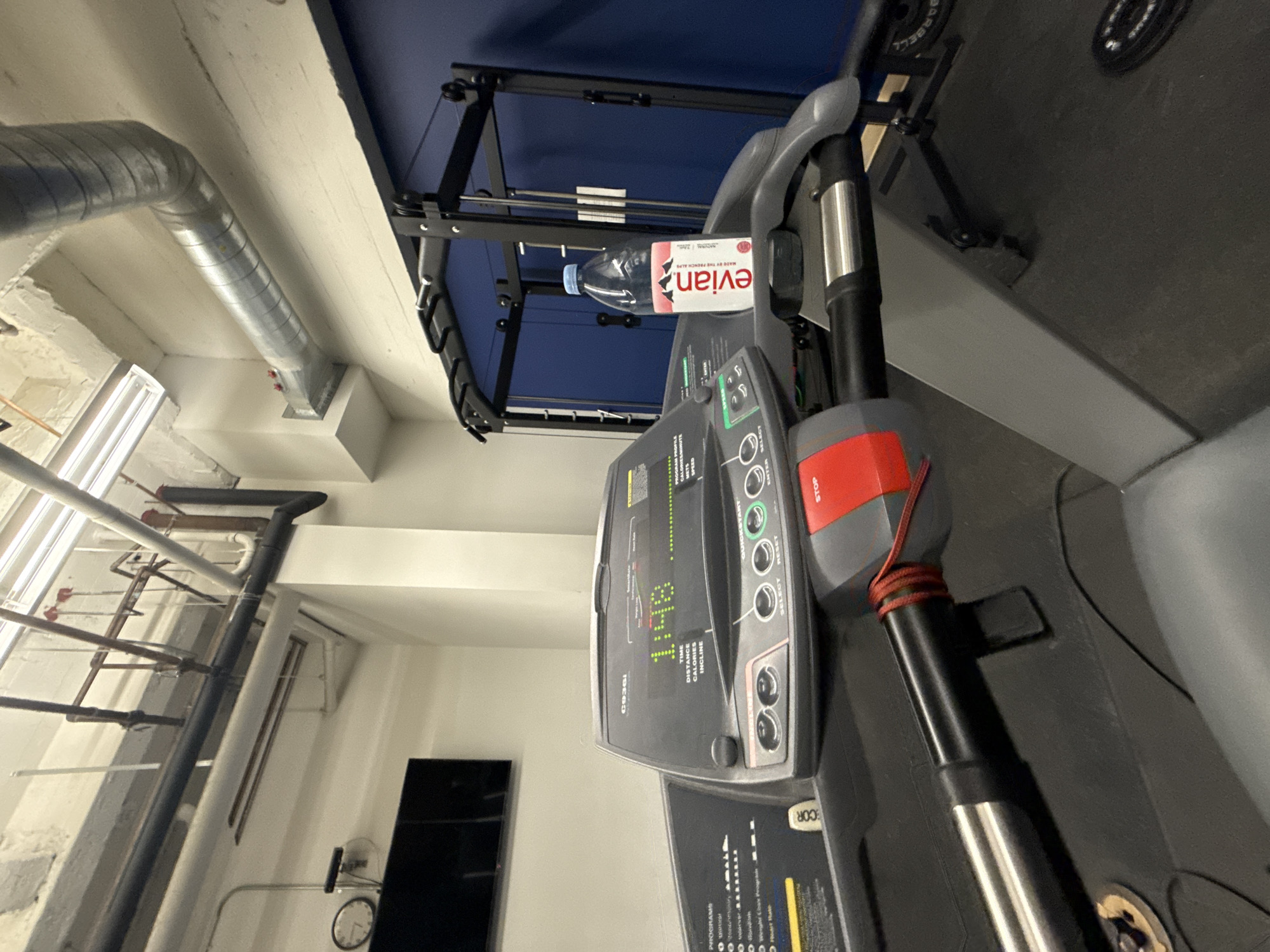 The image presents a partially visible treadmill, positioned in a room setting. Notable features on the treadmill include a control panel displaying metrics and the brand name 'COR'. A bottle of Evian water rests on the treadmill's support structure.
The broader environment includes an industrial-style ceiling with exposed pipes and fixtures. A television screen can be seen partially in the background. The image's exact location within Los Angeles, United States of America, cannot be determined based on the visual information provided.