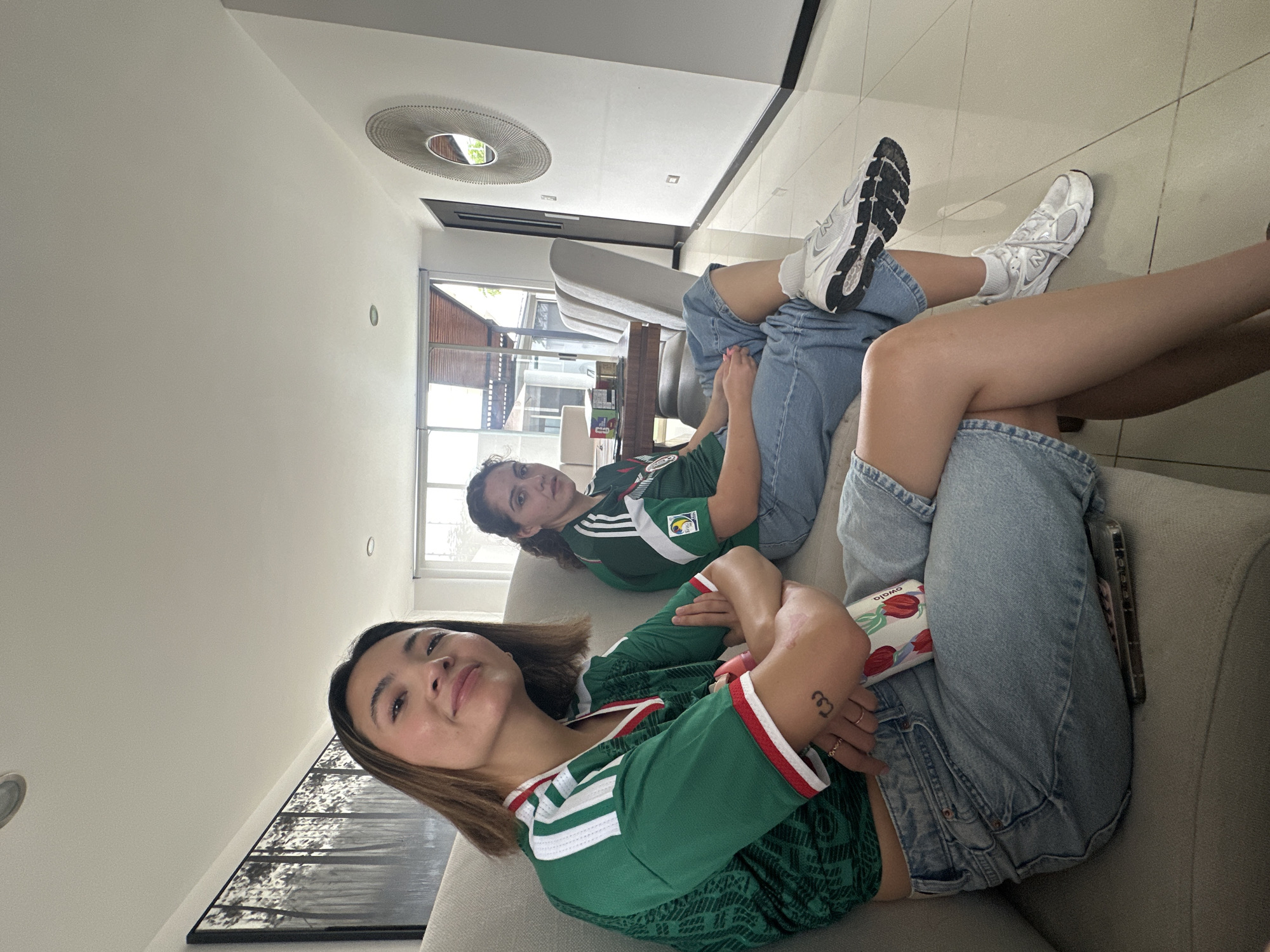 Two young women are seated on a light gray sofa in what appears to be a modern living room. The woman in the foreground is wearing a green and white striped soccer jersey, denim shorts, and white athletic shoes. She has her arms crossed and is looking towards the camera with a slight smile. A small, pink object and a phone are on the sofa near her. The woman in the background is wearing a similar green soccer jersey and blue jeans, and is looking off to the side. Behind them, a wooden dining table with chairs is visible, along with a large abstract artwork on the wall and a decorative mirror. The floor is tiled, and large windows with wooden blinds are present in the background, suggesting a bright, daytime scene. The overall impression is of a casual gathering in a well-lit residential interior, possibly in San Bernardino Tlaxcalancingo, Mexico, given the context provided.