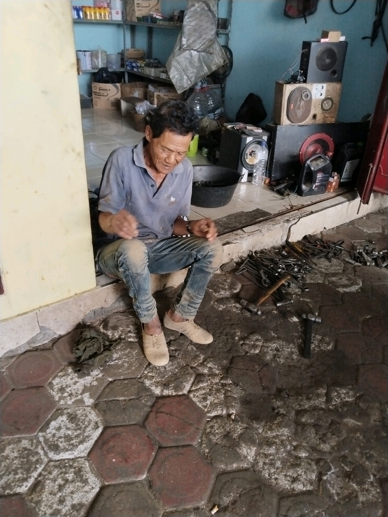 In a dimly lit workshop in Prigi, Indonesia, a man of East Asian descent, appearing to be in his late 50s or early 60s, sits on a concrete ledge. He is wearing a short-sleeved blue polo shirt and faded blue jeans, both appearing dusty. His feet are clad in light-colored, low-top sneakers.
The man's posture suggests he is resting. His hands are clasped in his lap, and he is looking downwards, possibly in thought or weariness. His facial features are weathered, with visible wrinkles.
The workshop environment is cluttered. To the man's right, a collection of various metal tools, including hammers and wrenches, are scattered on the ground. Beyond the tools, various objects are piled, including what appear to be speakers stacked on top of each other, plastic containers, and a large black basin. The floor is a mix of hexagonal paving stones and dirt. The walls are painted a light blue. The overall impression is of a functional, possibly a bit disorganized, workspace.
