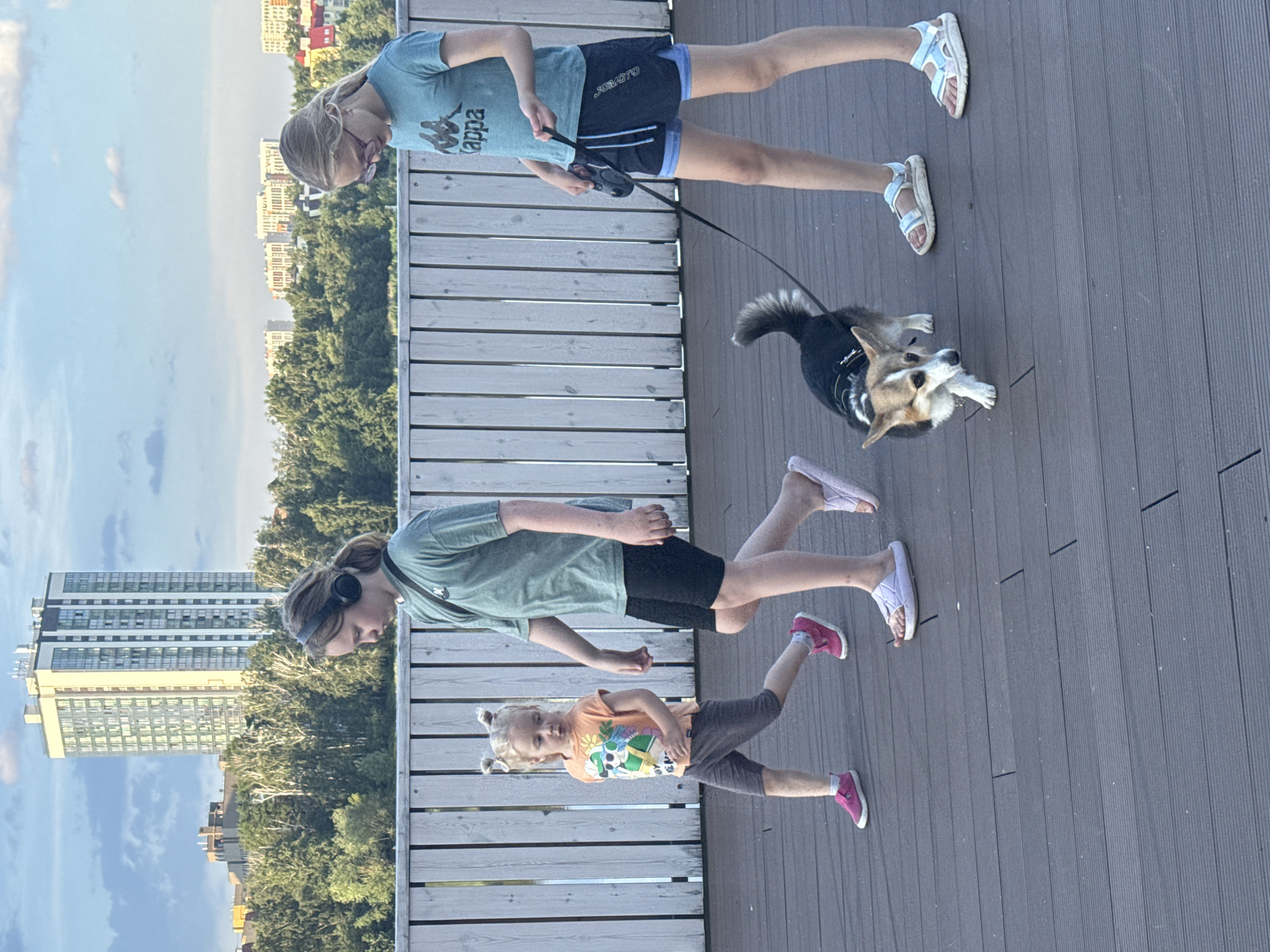 In Yekaterinburg, Russia, on a wooden walkway, three individuals and a dog are captured in a full-body, eye-level shot. The scene is set against a backdrop of a cityscape with tall buildings and lush green trees under a partly cloudy sky.
On the left, a young girl with blonde hair styled in two pigtails, wearing an orange t-shirt with a graphic print and dark grey shorts, walks with pink sneakers and white socks. To her right, a person with brown hair, wearing headphones, a green t-shirt, and black shorts, walks with a purposeful stride. In the center, a small corgi dog, with brown and white fur, is on a leash, looking to its left. To the right, a person with blonde hair and glasses, wearing a light blue t-shirt and dark blue shorts over a lighter blue layer, walks the dog, holding the leash with their right hand. This person is wearing light blue and white sandals. The walkway is made of weathered grey wooden planks.