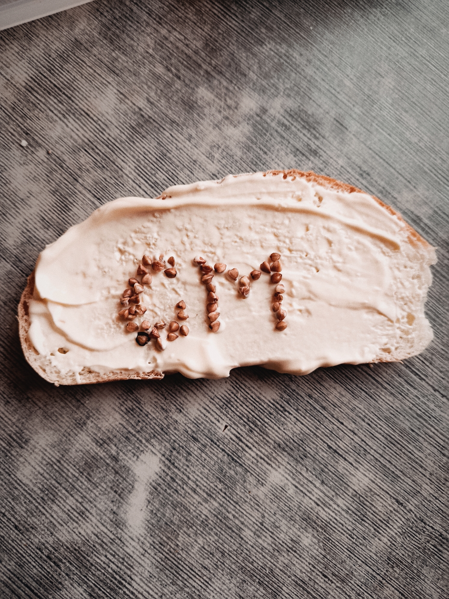 A slice of bread topped with a creamy spread is seen on a wooden surface. The letters "GM" are spelled out using small brown grains on the spread. The environment is not distinct enough to identify the city of Фоки, Russia.