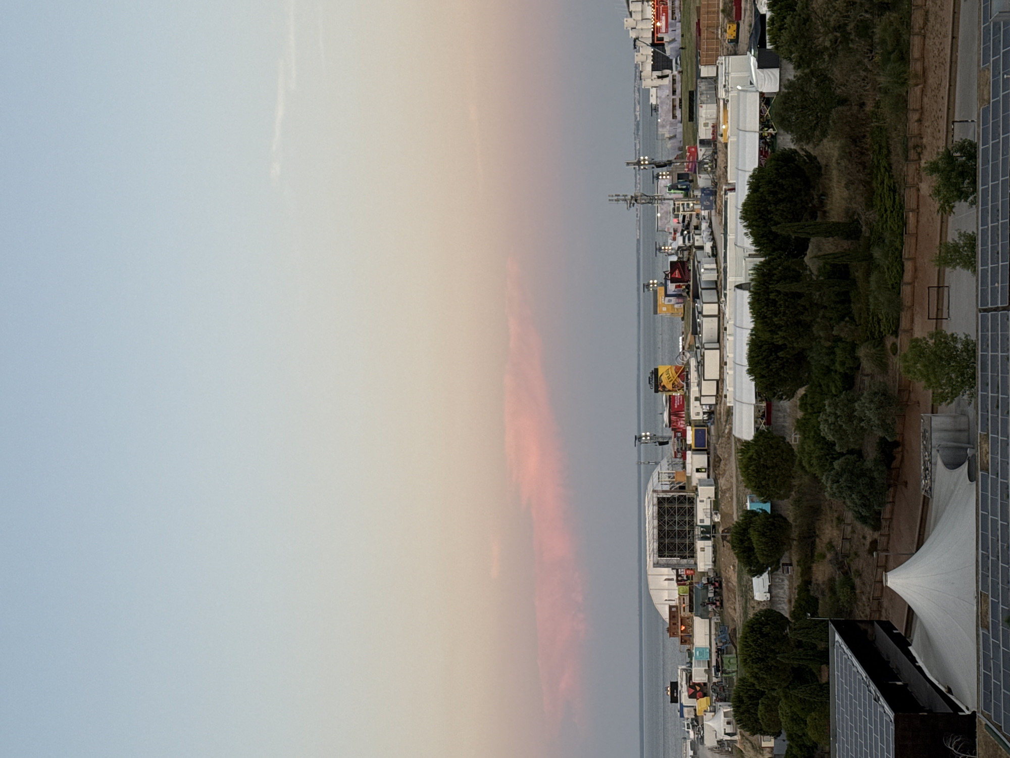 The image depicts an aerial view of a festival or event setup near the Tagus River in Loures, Portugal, during twilight. The sky displays soft hues of blue and pink from the setting sun. In the mid-ground, a large construction site is visible, featuring temporary structures, white modular units, and scaffolding for stages or support. Several trucks and vehicles are parked among the structures, indicating ongoing preparation or activity. A prominent white tent structure with a conical roof is situated in the foreground, adjacent to a building with solar panels. The background reveals the expanse of the Tagus River and distant, hazy shorelines. The overall scene is one of a large-scale event in its early stages of development or breakdown, set against a coastal urban or semi-urban landscape.