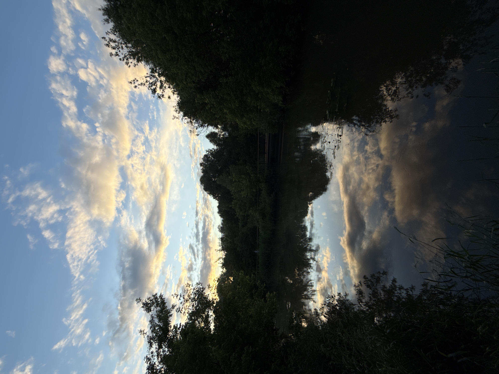 The image depicts a tranquil river scene at dusk, with the sky a canvas of blues and whites, illuminated by the warm glow of the setting sun reflecting on the clouds. Lush green trees line the banks of the river, their dense foliage creating a natural frame for the water. A small bridge is visible in the distance, spanning the river, and a solitary figure can be seen walking across it. The water's surface is calm, mirroring the sky and trees, creating a sense of serene reflection. The overall impression is one of peace and natural beauty, with the subtle hints of human presence adding a touch of understated life to the otherwise untouched landscape.