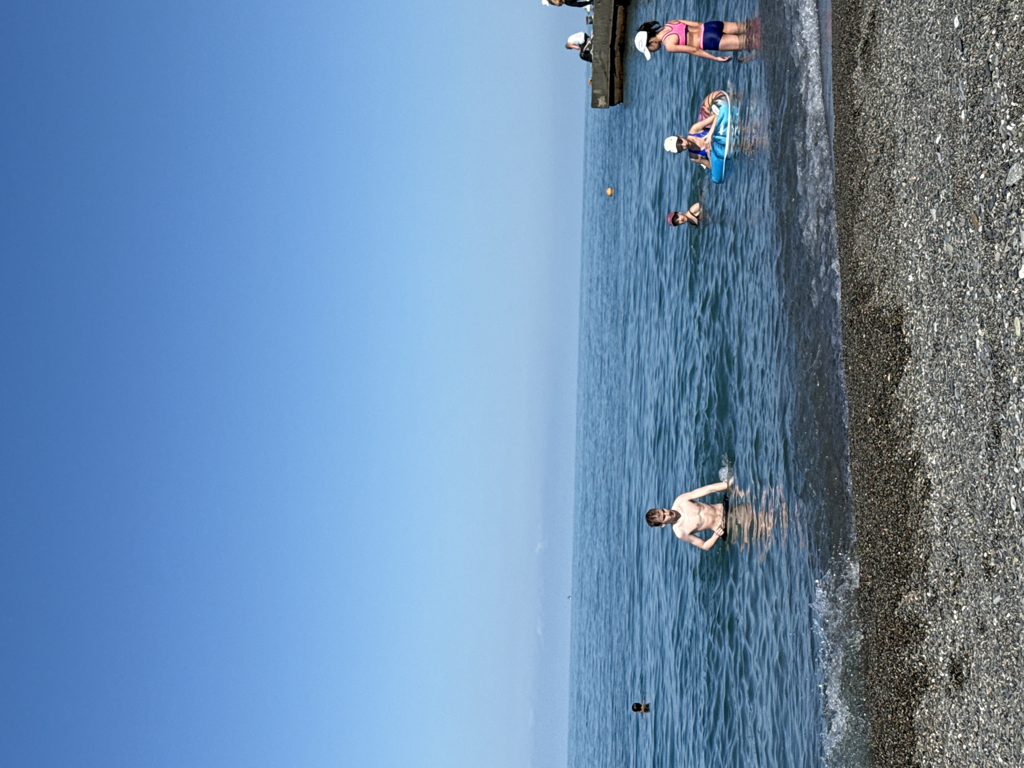 The image depicts a sunny day at a pebble beach in Sochi, Russia. A clear blue sky occupies the left side of the frame, contrasting with the tranquil blue water of the sea on the right. Several people are enjoying the water and the beach.
In the sea, a man with a beard is partially submerged, looking towards the camera with a relaxed expression. Further out, a person is on a colorful inflatable ring, and another person in a blue swimsuit is visible nearby. Closer to the shore, a person wearing a pink bikini and a white hat is standing.
The beach itself is covered in dark grey pebbles. Waves gently lap at the shore, indicating calm weather. The overall scene suggests a pleasant summer day at the seaside, with people engaging in typical beach activities like swimming and relaxing. There is no visible text in the image.
