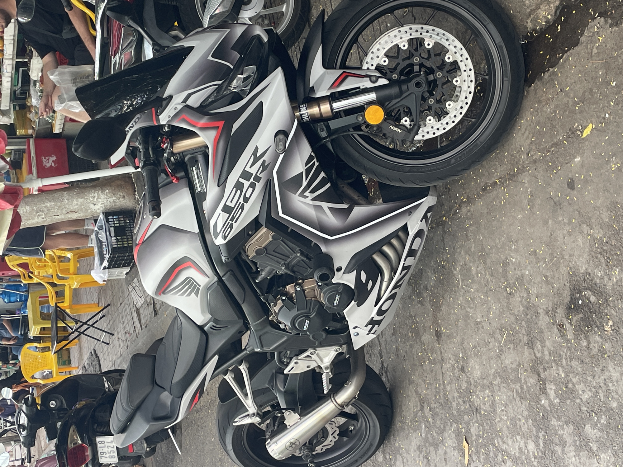 The image shows a Honda CBR650R motorcycle parked on a paved street, likely in a city environment. The motorcycle is predominantly gray and black with red accents and features the "CBR" and "HONDA" logos. It is positioned diagonally in the frame, with the front wheel facing towards the right and the rear wheel towards the left. The background shows glimpses of a street scene with a black scooter parked to the left of the motorcycle, yellow plastic chairs and a small black table on the right, and what appears to be a market stall with various items and people in the distance. The ground is a textured gray pavement with scattered small yellow debris, possibly fallen petals or leaves. The lighting suggests it is daytime, with shadows cast by the objects. The scene depicts a common urban setting where motorcycles are a prevalent mode of transportation.