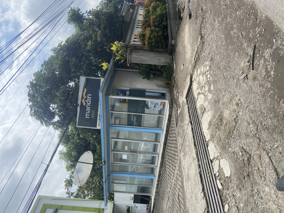 A small, blue and white building, labeled “Mandiri ATM”, sits in front of a large, mature, green leafy tree, in the city of Ciparay, Indonesia. The building has a glass facade with large, frosted glass panels. Inside, the building houses an ATM machine. The building sits on a cracked gray concrete sidewalk. A satellite dish is mounted on the roof of the building. There are power lines running overhead and a large, green leafy tree in the background. The scene is captured from the perspective of a person standing on the sidewalk in front of the building. The scene is captured during the day in daylight. The building is located at a crossroads. There are no people present. The scene is a typical example of a street corner in a developing country.