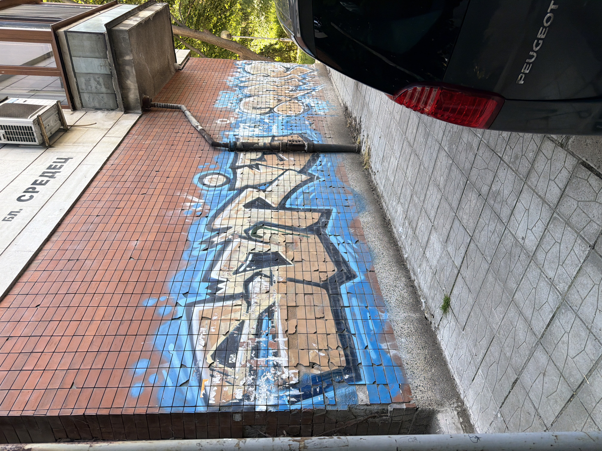 The image depicts a section of a brick building wall covered in blue and tan graffiti. The graffiti appears to be stylized lettering, with some parts peeling away from the wall, revealing the underlying brick. A drainpipe runs vertically down the wall. Above the graffiti, the building has a white facade with black lettering that reads "БЛ. СРЕДЕЦ". An air conditioning unit is mounted on the white facade. To the right, a portion of a dark-colored Peugeot car is visible, with its taillight in the foreground. The ground is paved with gray concrete tiles arranged in a geometric pattern. A small clump of grass is growing between the tiles. The overall scene suggests an urban environment.