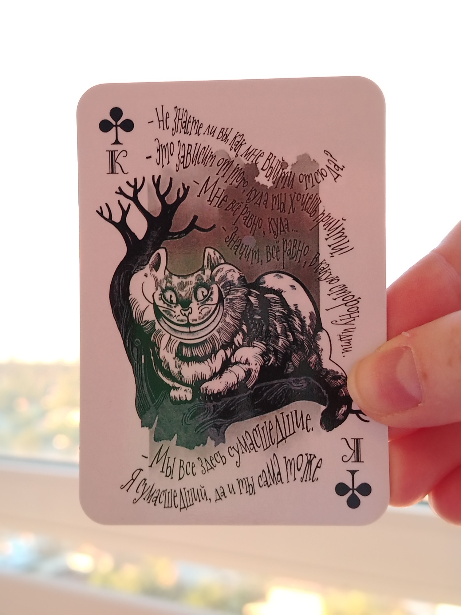 A hand holds up a decorative playing card featuring a stylized illustration of the Cheshire Cat from "Alice in Wonderland." The card, which appears to be a King of Clubs (К♣), is held against a blurred background showing soft, warm light, suggesting an indoor setting near a window during daytime, possibly late afternoon. The scene takes place in Izhevsk, Russia.
The card's design includes the Cheshire Cat perched on a bare tree branch, grinning widely. The card's background is a light pinkish-beige. Black text in a whimsical, handwritten font is printed on the card, quoting famous lines from the story in Russian.
The text on the card reads:
"- Не знаете ли вы, как мне выйти отсюда. (Do you know which way I ought to go from here?)"
"К - Это зависит от того, куда ты хочешь прийти! (That depends a good deal on where you want to get to!)"
"- Мне всё равно, куда... (I don't much care where...)"
"- Значит, всё равно, в какую сторону идти. (Then it doesn't matter which way you go.)"
"- Мы все здесь сумасшедшие. (We're all mad here.)"
"Я сумасшедший, да и ты сама тоже. (I'm mad. You're mad. And you yourself too.)"
The card combines classic literature with a playing card motif, making it a unique item.