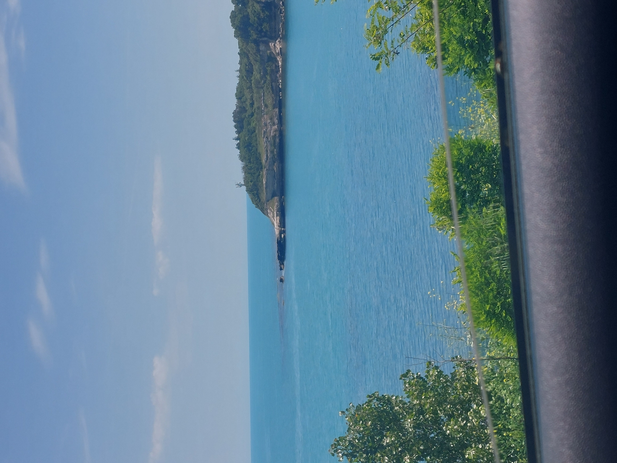 The image captures a view of a body of water, likely a sea or lake, with a promontory or cliff covered in greenery extending into it. The water is a bright blue color. The sky is light blue with some wispy clouds. Some green trees and foliage are visible in the foreground, partially obstructing the view. A dark vertical element on the right side of the image seems to be part of the frame, perhaps a window or door jamb. It is not possible to determine if the location is in Tsarevo, Bulgaria from the visual information alone.