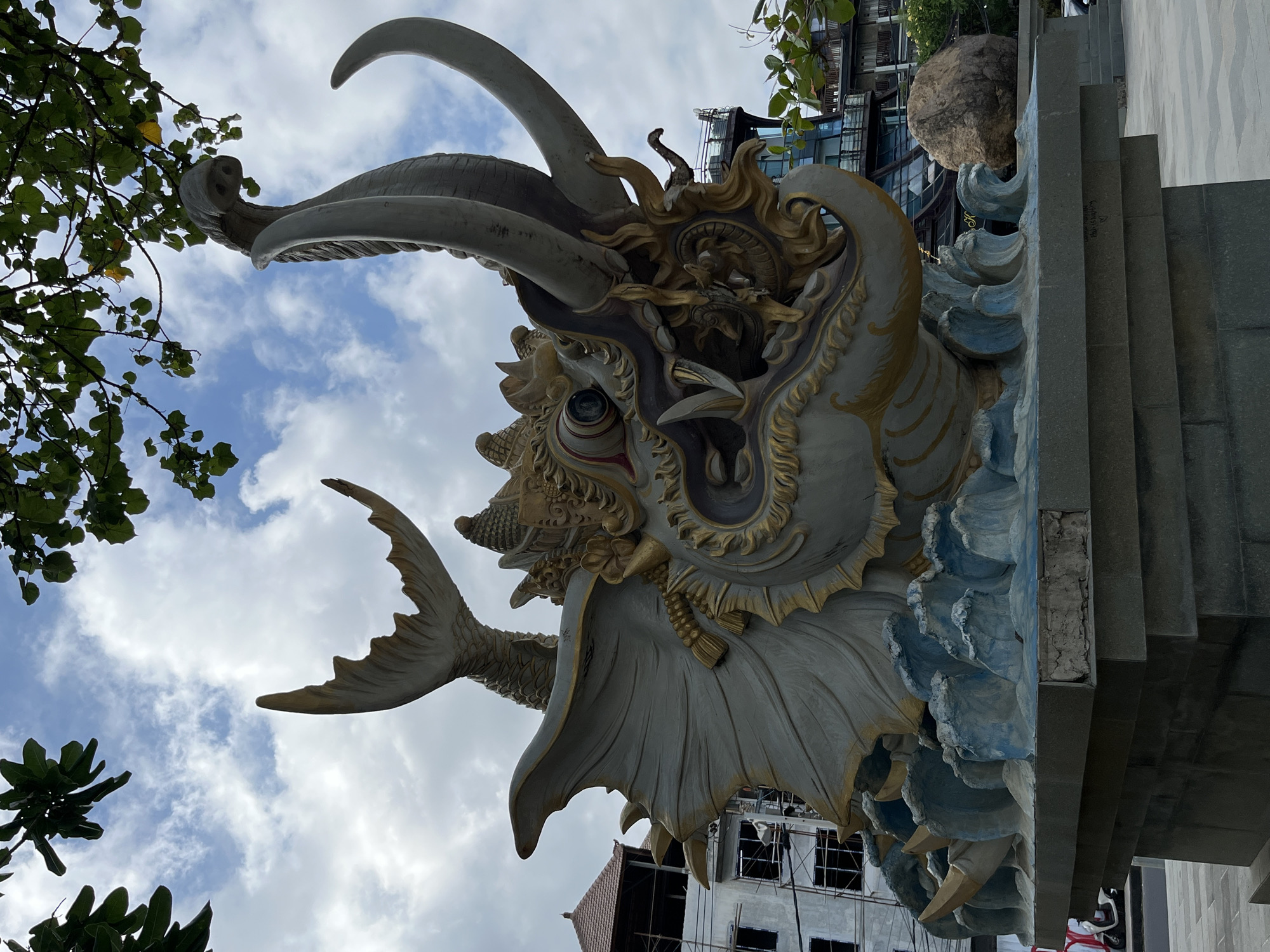 This media file depicts a close-up, low-angle shot of a large, ornate dragon-like sculpture against a cloudy sky. The sculpture, rendered in shades of grey, white, and gold, features intricate details such as scales, fangs, large horns, and a finned tail. The dragon's head is the primary focus, with its mouth agape and eyes wide open, giving it a fierce and dynamic appearance. The sculpture is adorned with golden accents that highlight its mythological features.
Part of a building under construction, with scaffolding and visible concrete structures, is visible in the background, suggesting an ongoing development or renovation project. Lush green foliage from trees frames the top left of the image. The sky is filled with white, puffy clouds, indicating fair weather, possibly during the daytime. The sculpture appears to be a decorative element, possibly part of an entrance or a landmark in Kuta, Indonesia, a location known for its vibrant culture and architecture. The upward perspective emphasizes the scale and grandeur of the artwork. There is no visible text in the media file.