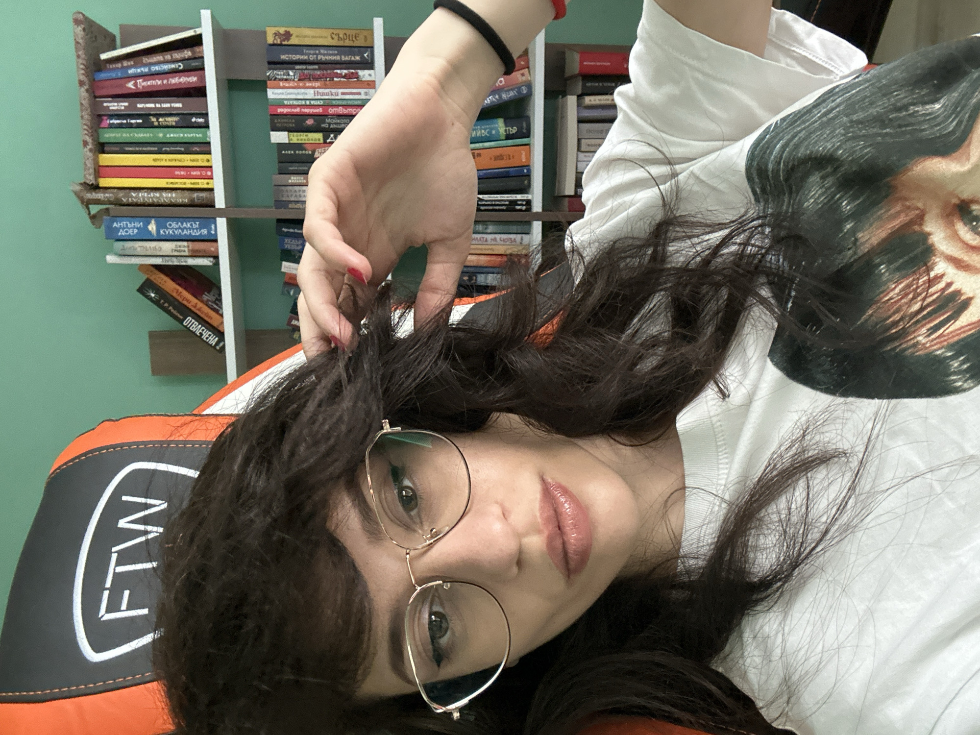 This is a portrait of a young woman with dark, wavy hair, wearing a white t-shirt with a graphic print of a face. She has on round, gold-rimmed glasses and her lips are painted with a nude lipstick. She is lying down and her left hand is reaching up to touch her hair, with bright red nail polish visible on her fingernails. A black band is on her wrist, along with a red string bracelet.
The background shows bookshelves filled with books in Bulgarian. The spines of several books are visible, with titles such as "СЪРЦЕ", "ИСТОРИИ ОТ РЪЧНИЯ БАГАЖ", and "ОТВЛЕЧЕНА". The woman is resting on an orange and black chair with "FTW" written in white letters. The lighting suggests it could be daytime, and the setting appears to be indoors, likely a room with a green wall. The overall mood is casual and relaxed.