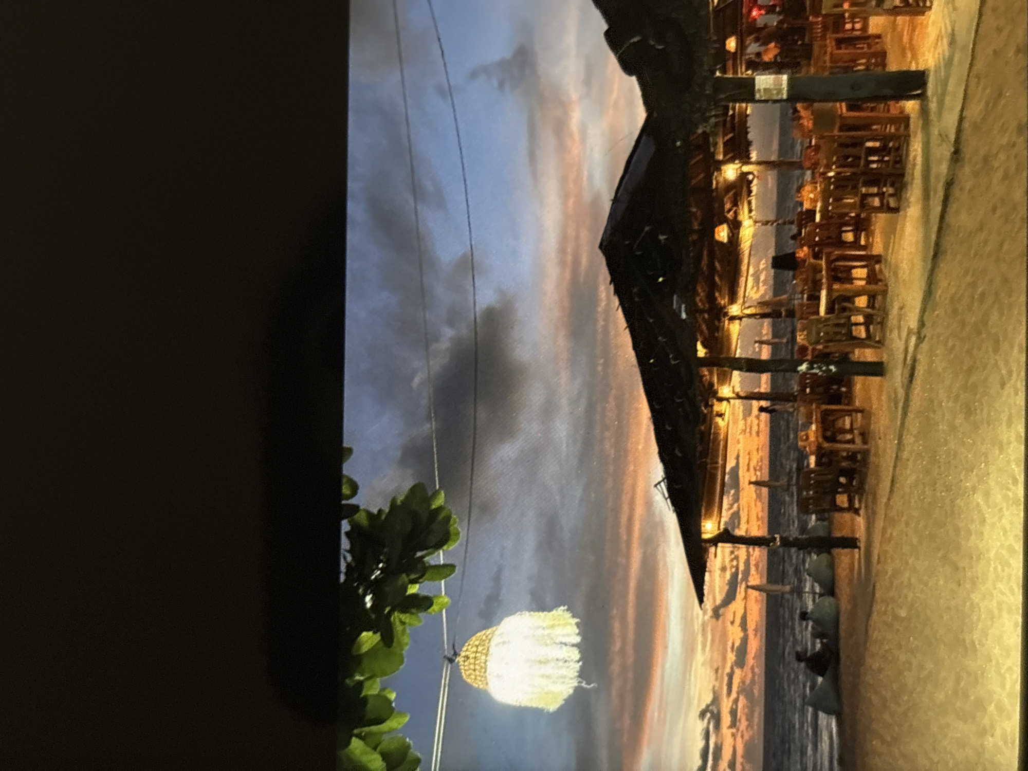 This is a wide shot of a beachside restaurant at dusk in Chalong, Thailand. The sky displays a gradient of blues and oranges as the sun sets over the ocean. Palm trees and string lights are visible, and a hammock hangs in the foreground. Tables and chairs are set up on the sand, indicating an outdoor dining area. The scene suggests a relaxed and tropical atmosphere.